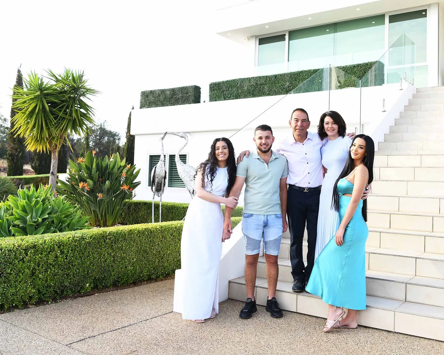 Group of five people standing on outdoor stairs, smiling, with a modern white building, lush green plants, and two decorative herons in the background.