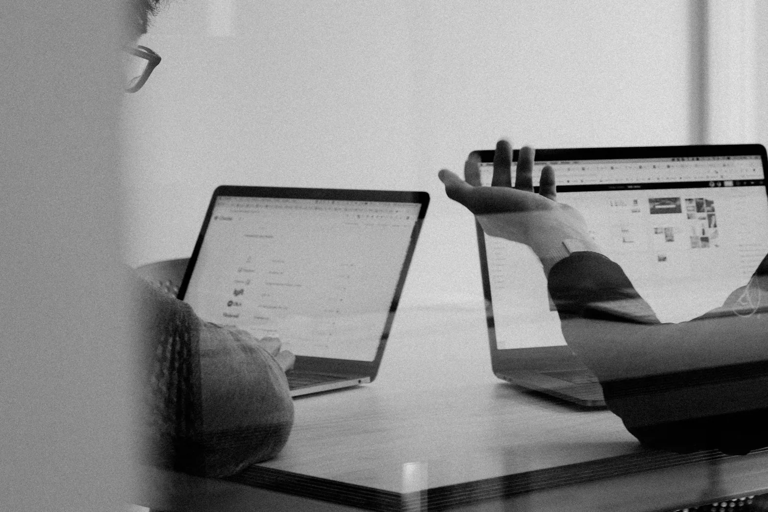 Two people using laptops at a table, one person is reaching towards the screen; the image is in black and white.