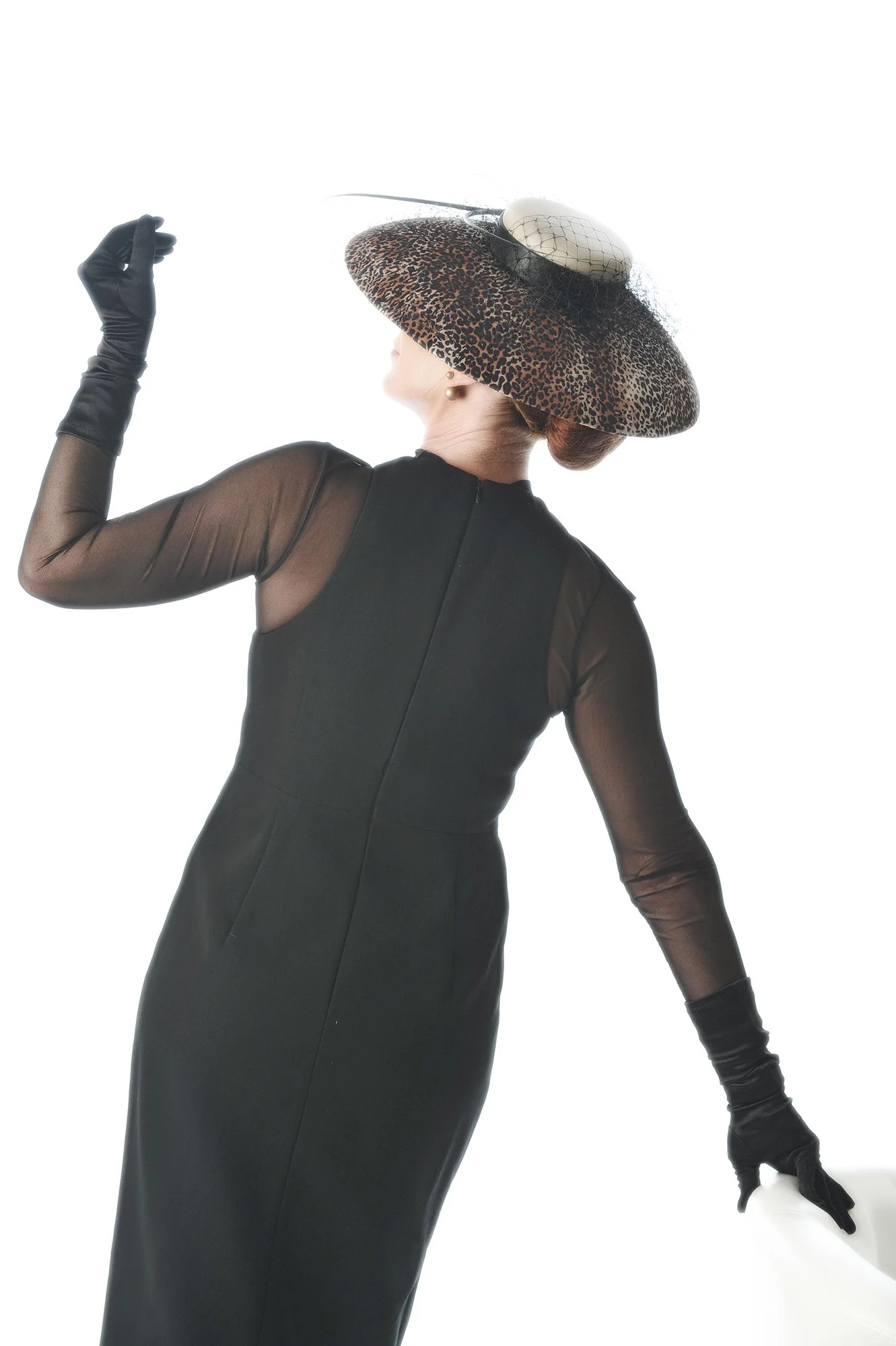 Woman in black dress with sheer sleeves and leopard-print wide-brim hat with white hat and black veil, wearing black gloves, posing against a white background.