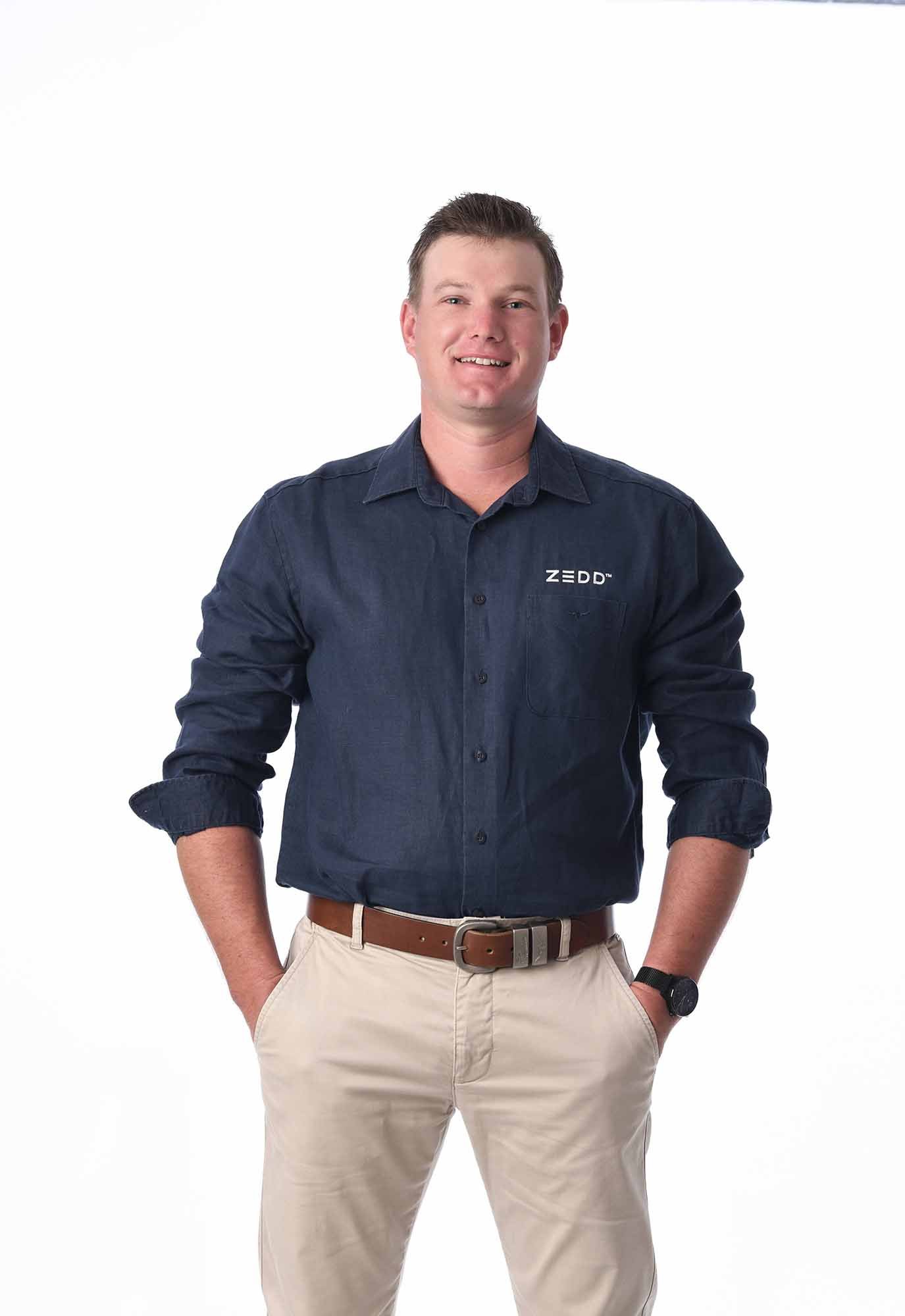 A young Caucasian man with short dark hair, wearing a navy blue button-up shirt with the logo 'ZEDD' on the chest, khaki pants, a brown belt, and a wristwatch, stands against a white background with his hands in his pockets, smiling at the camera.
