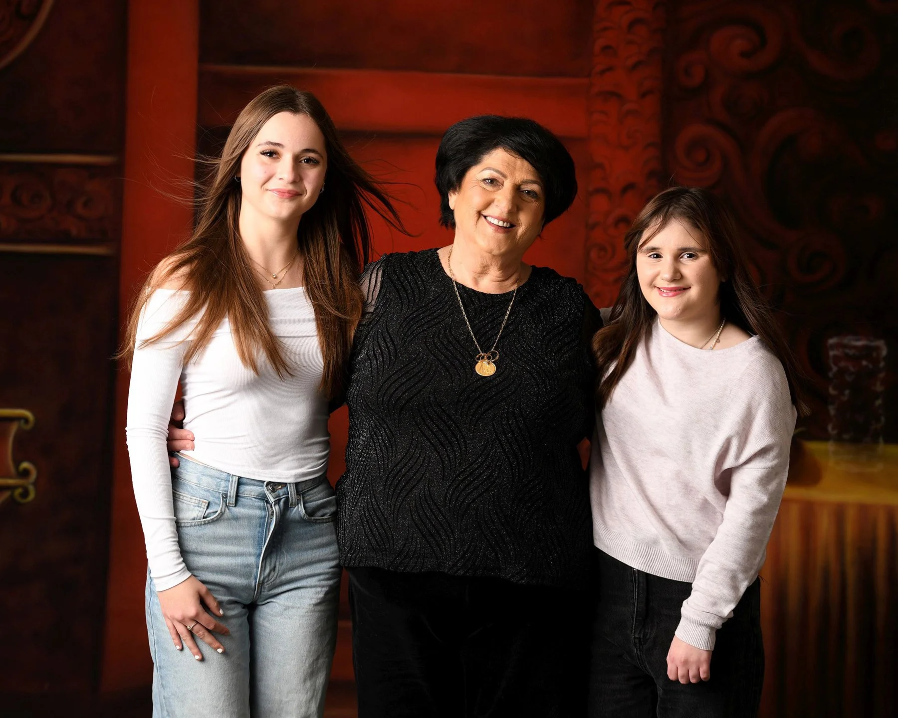 Three women standing close together, smiling at the camera indoors, with a red patterned background.