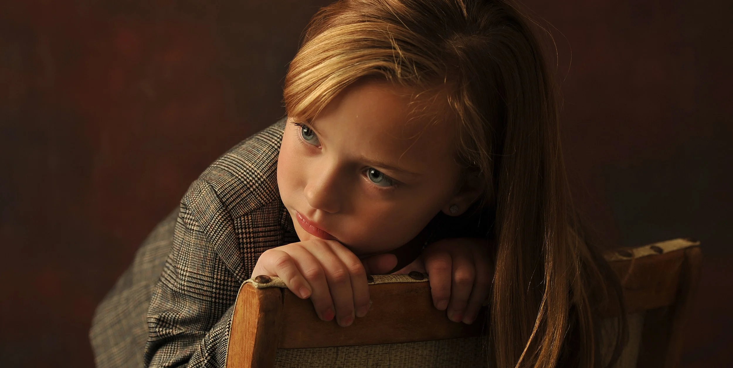 A young girl with long light brown hair and blue eyes, wearing a checkered blazer, leaning on a wooden chair with her chin resting on her hands, looking thoughtfully to the side against a dark brown background.