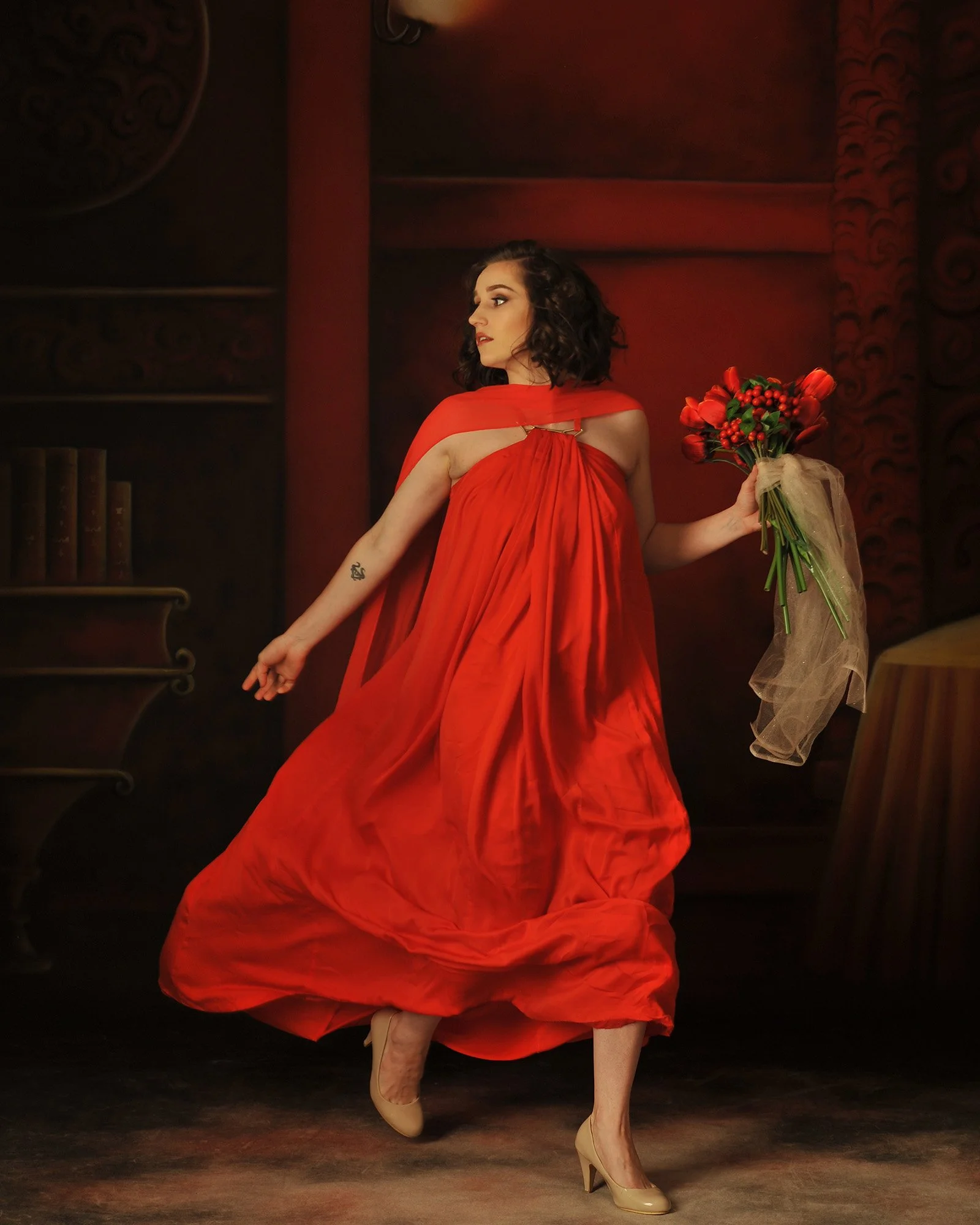 A woman in a flowing red dress holding a bouquet of red and orange flowers, standing indoors with dark walls and furniture.