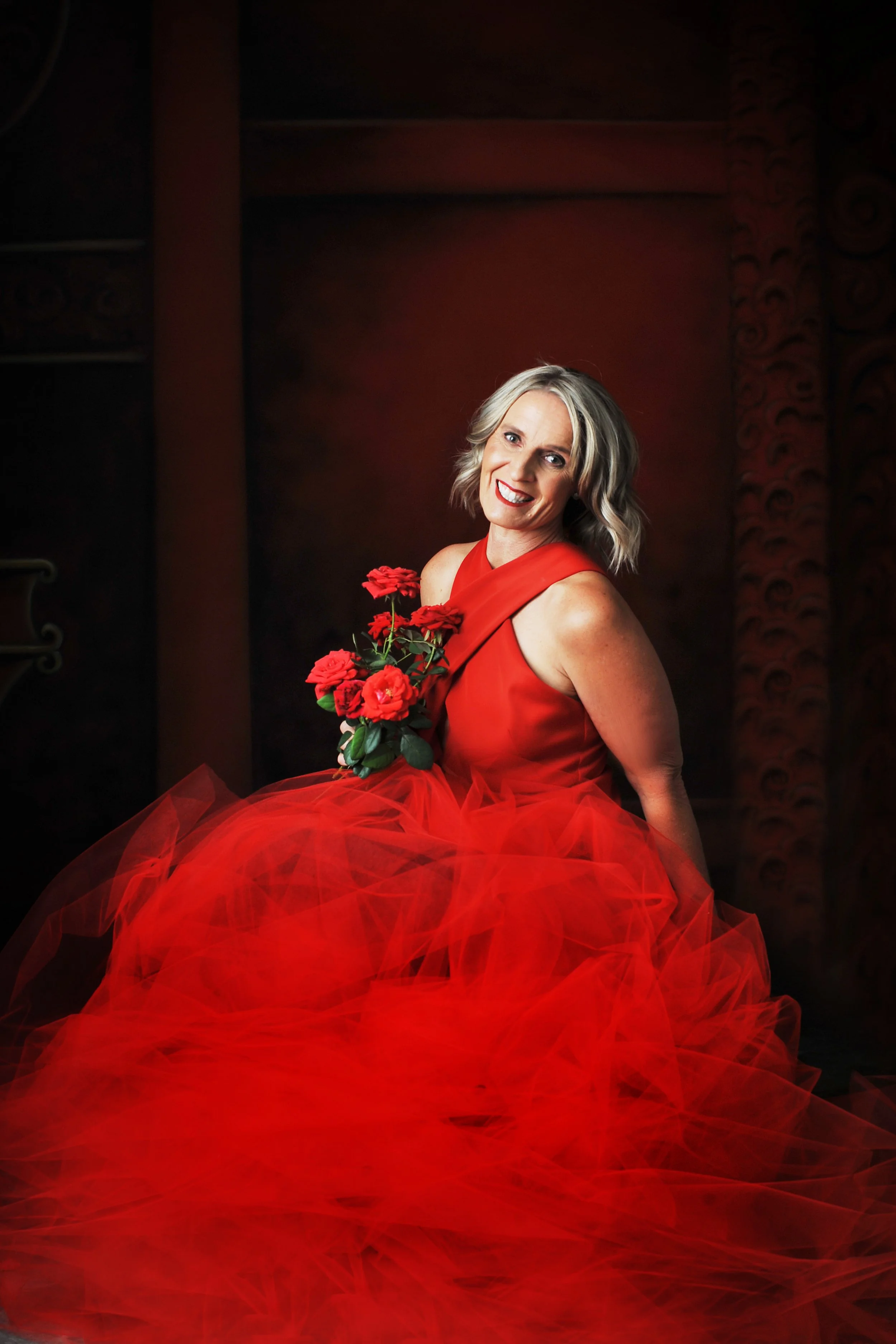 A woman with blonde hair smiling and wearing a red dress, holding a bouquet of red roses, posing against a dark, textured background.