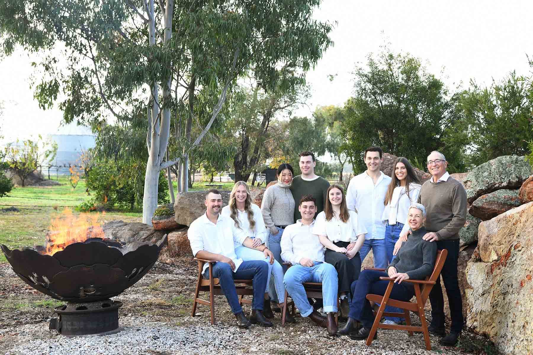 A group of eleven people, including adults and children, smiling and posing outdoors at sunset near a fire pit with flames, surrounded by rocks and greenery in a natural setting.