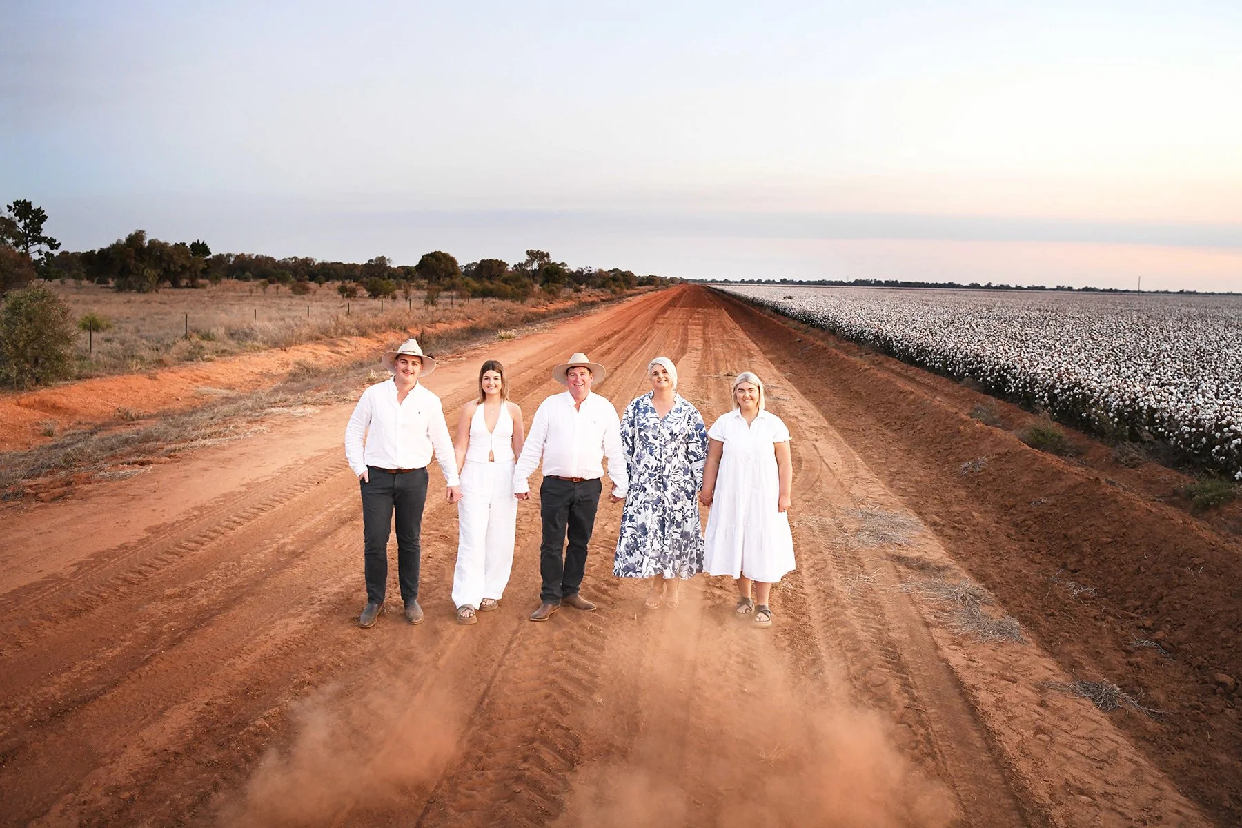 Group of five people walking hand in hand on a dirt road in a rural area with fields on either side, one with cotton plants and the other with dry grass and scattered trees, during sunset or sunrise.