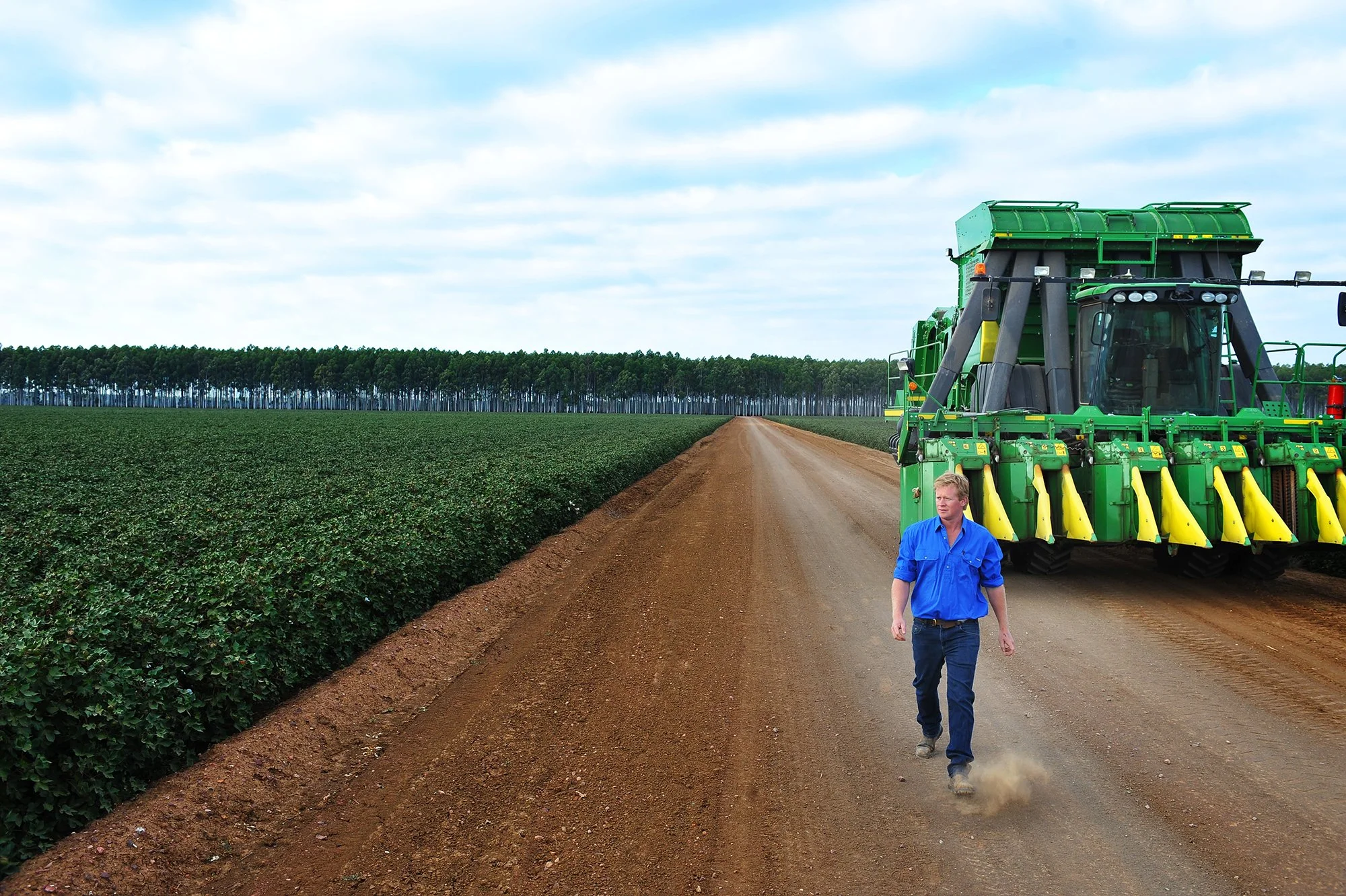 A farmer walking along a dirt road next to a large green tractor in a farm field with green plants. There are trees and a cloudy sky in the background.