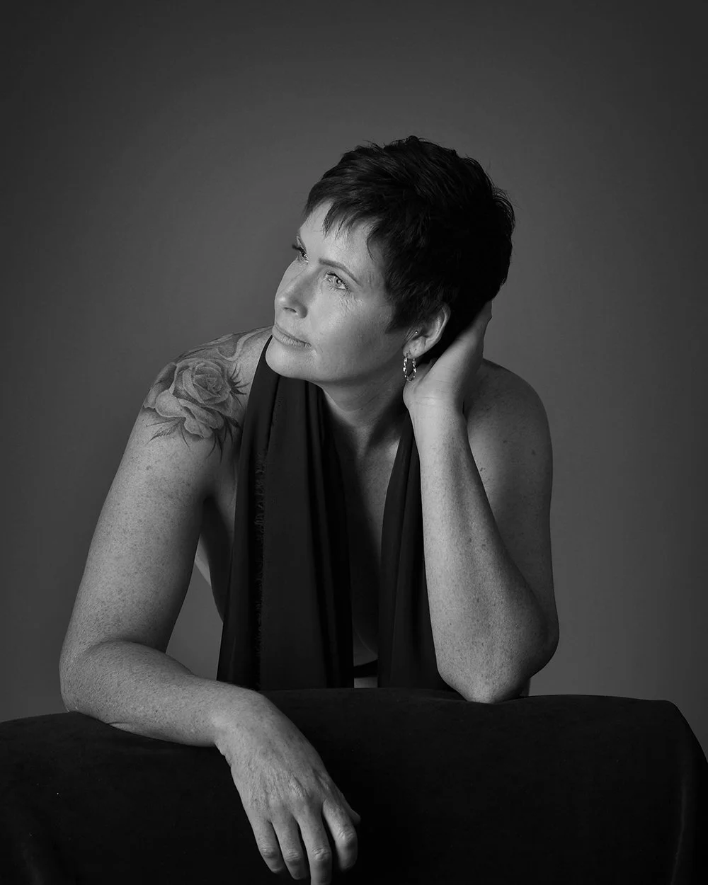 A black and white portrait of a woman with short dark hair, tattooed shoulder, earrings, resting her head on her hand, looking to the side, sitting at a table with a dark cloth.