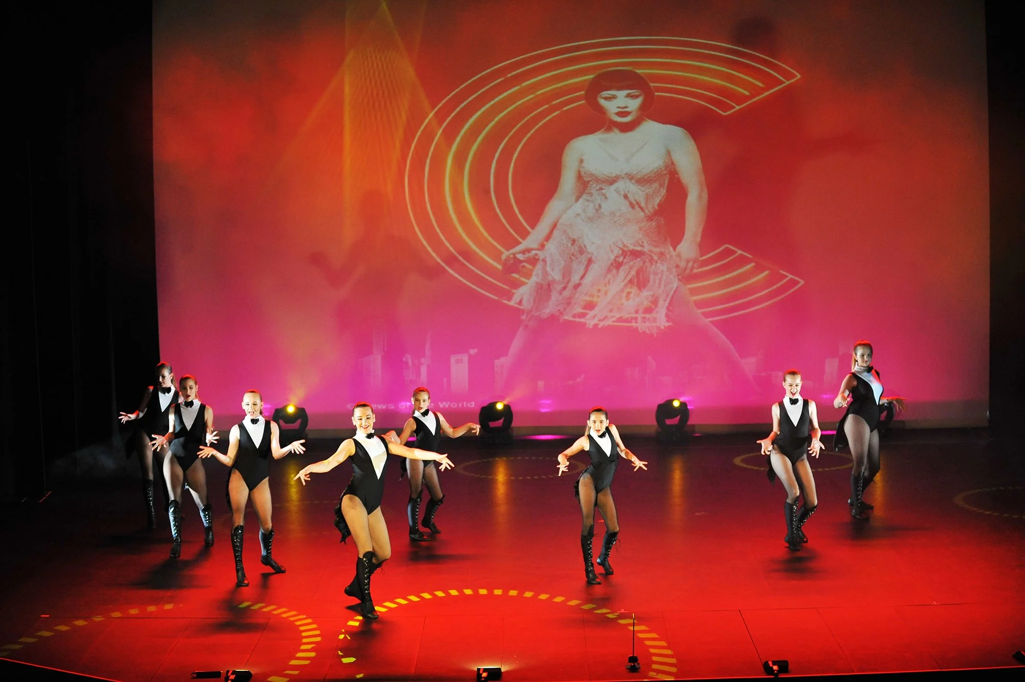Girls performing a dance routine on stage in black and white costumes with heels, with stage lighting and a large projected image of a woman in vintage dress behind them.