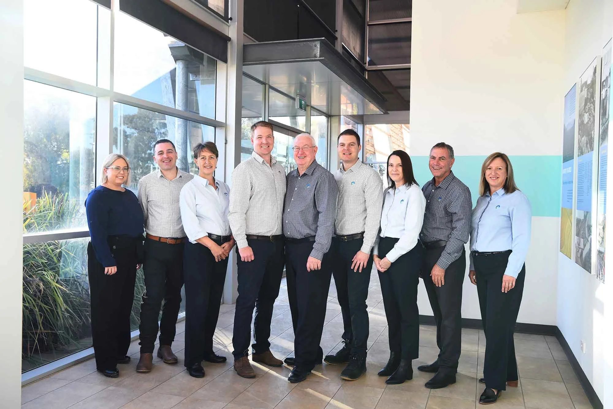 Group of nine professionals standing in a modern office lobby with large windows, smiling at the camera.