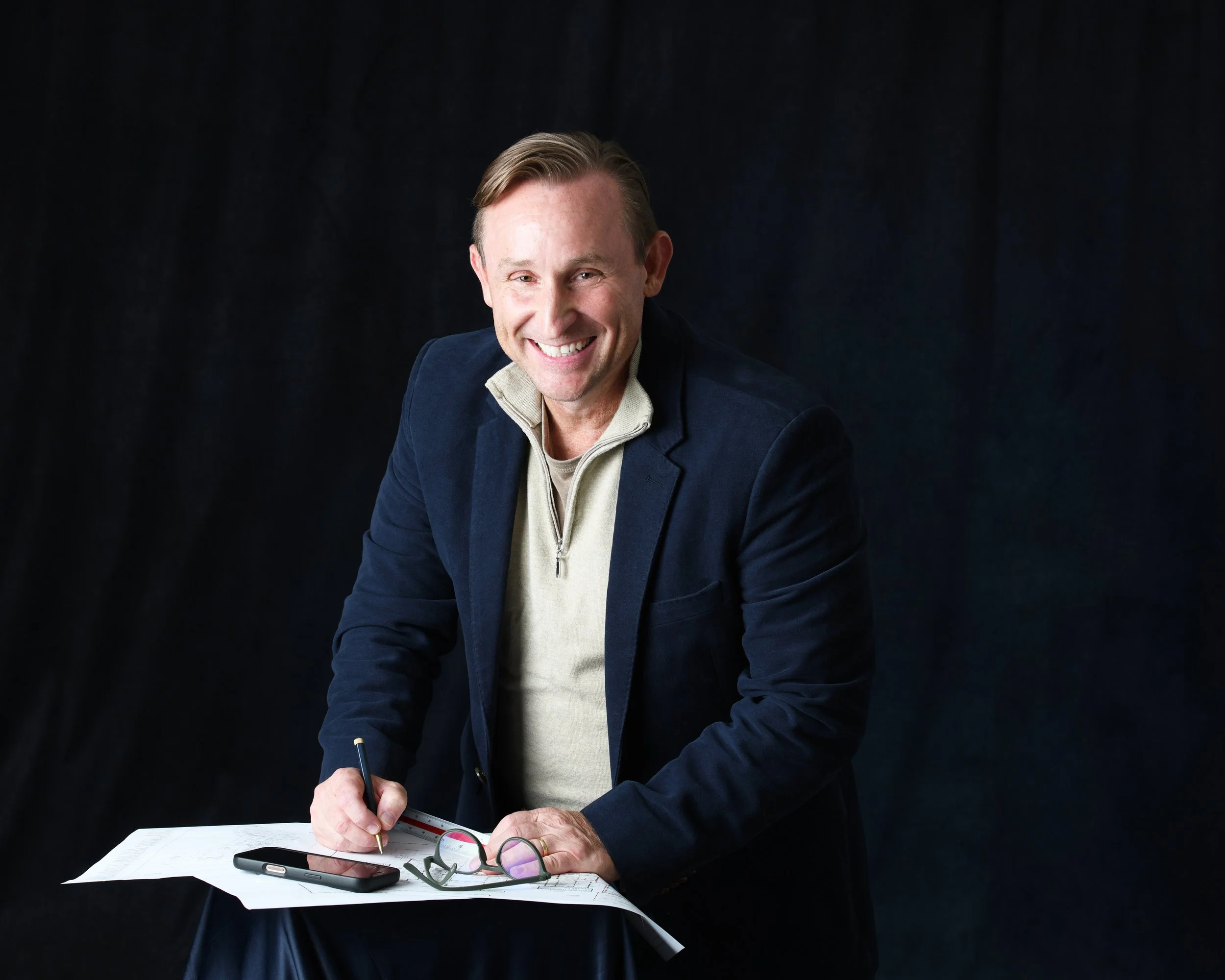 A man with light brown hair, wearing a dark blazer and a beige zip-up sweater, is smiling and sitting at a desk with various items including a pen, reading glasses, a smartphone, papers, and a calculator. The background is dark, highlighting the man 