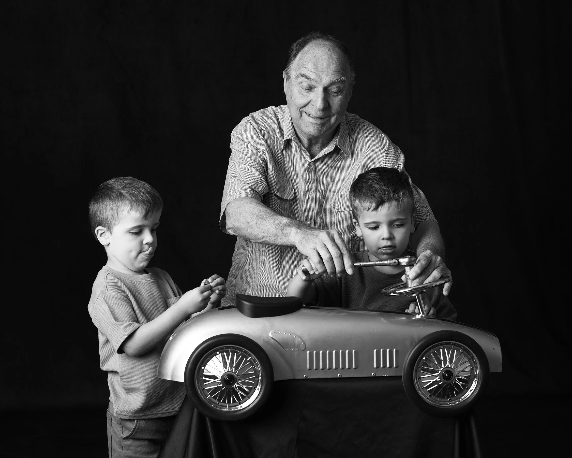 Black and white photo of an elderly man and two young boys sharing a moment over a toy car, with a dark background.