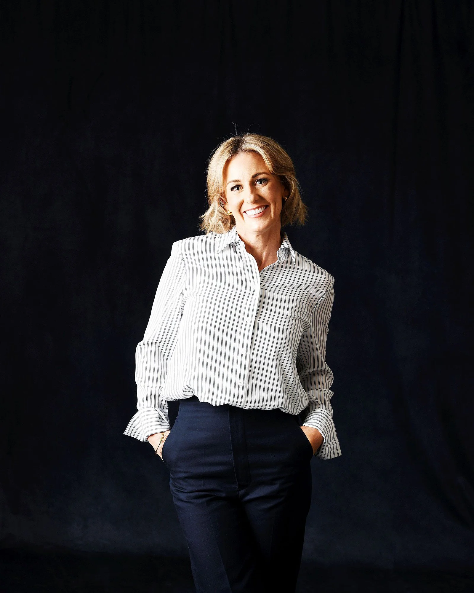 A woman with blonde hair in a striped professional shirt and dark pants, smiling with her hands in her pockets, standing against a black background.