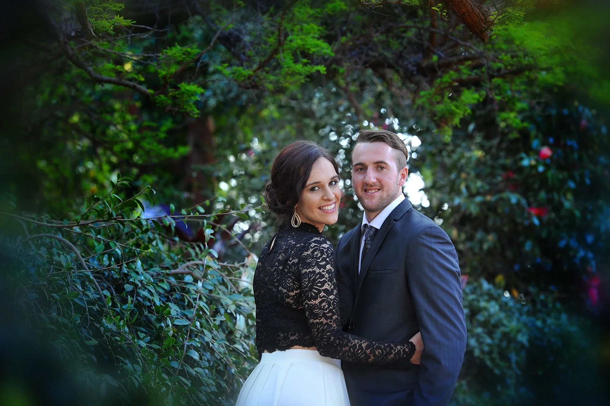 A happy couple, woman with dark hair in a lace black top and white skirt, and man with short hair in a dark suit, standing close outdoors among green trees and bushes, smiling at the camera.