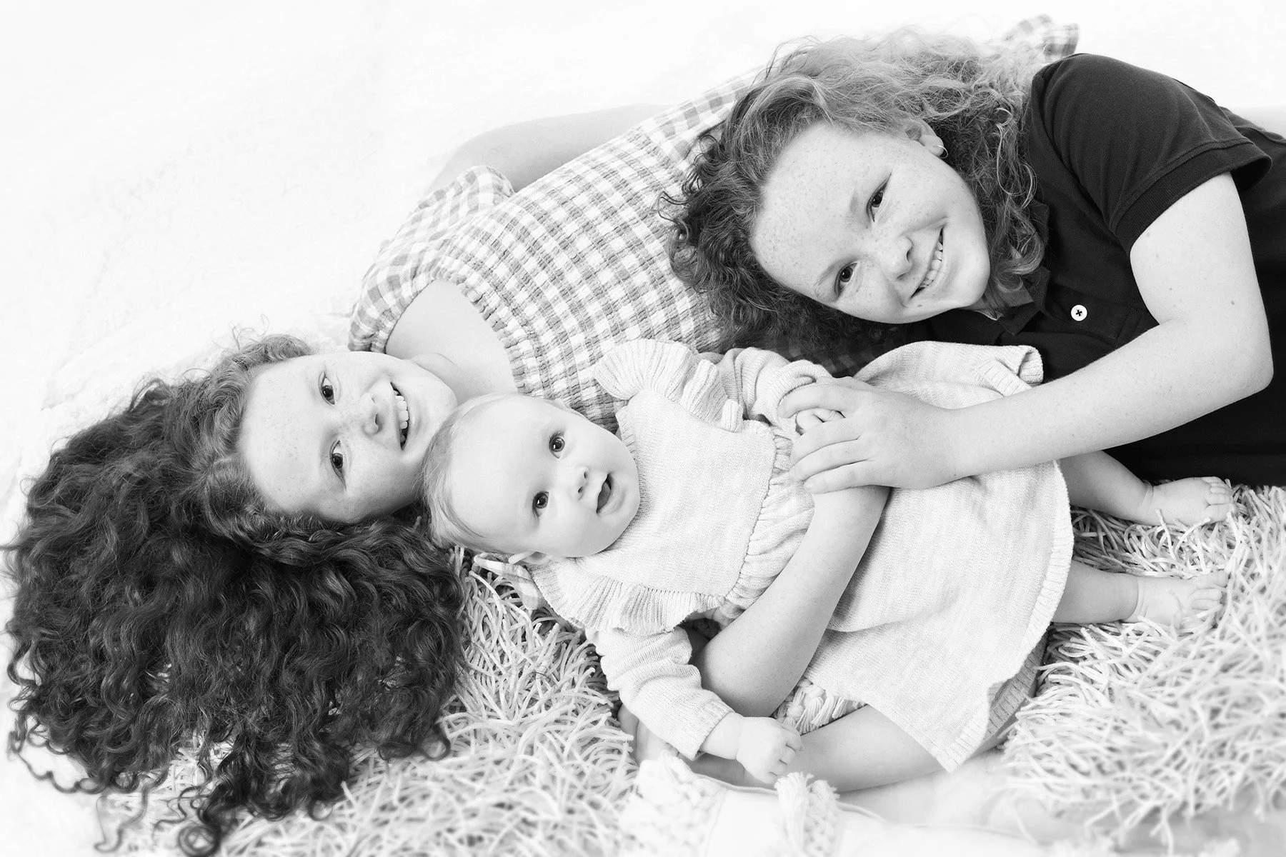 A black-and-white photo of three children lying on a bed with straw bedding. Two girls with curly hair and a young boy with straight hair are smiling and looking at the camera.