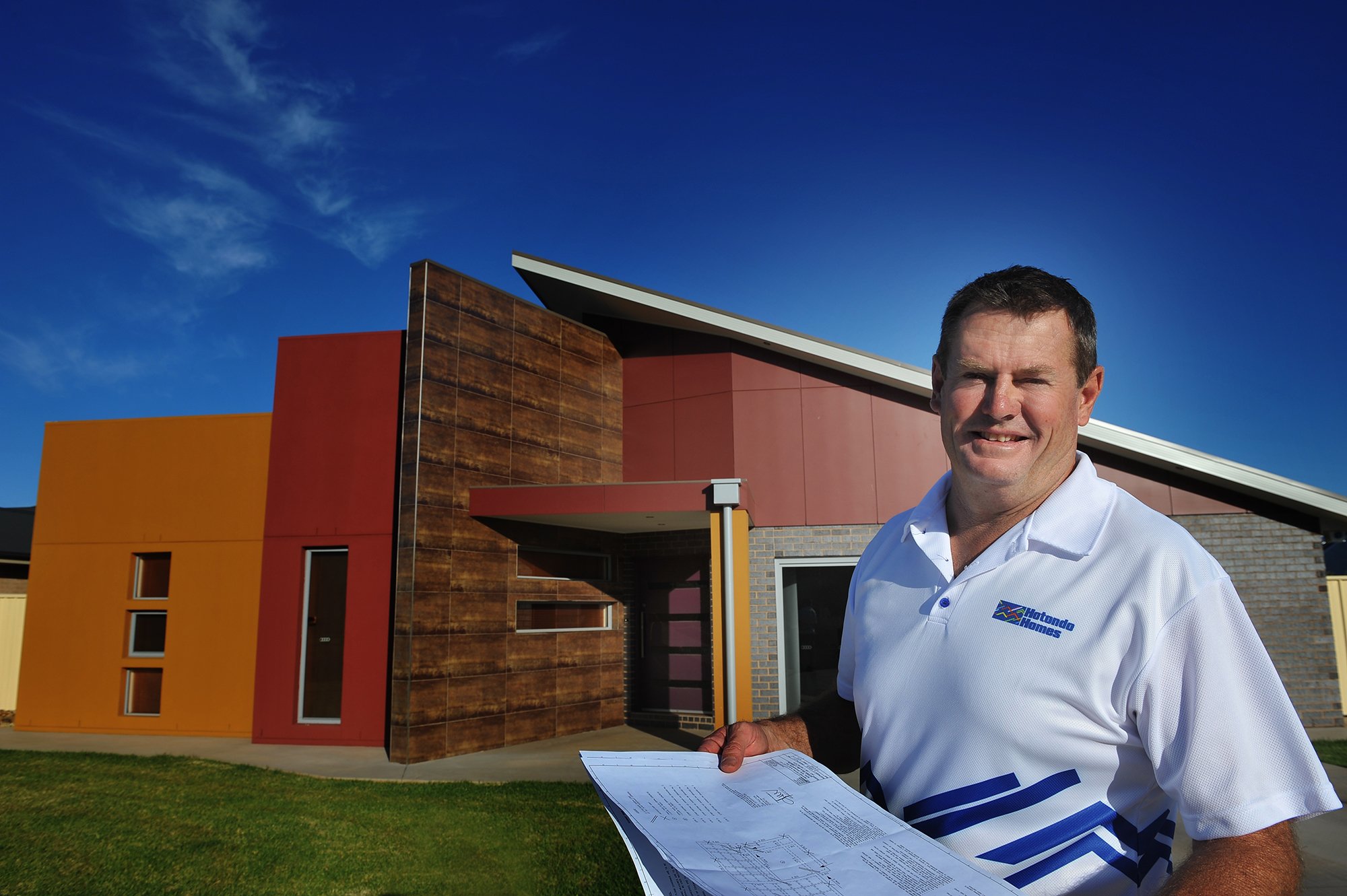 A man in a white polo shirt holding blueprints standing in front of a modern, colorful building on a sunny day.