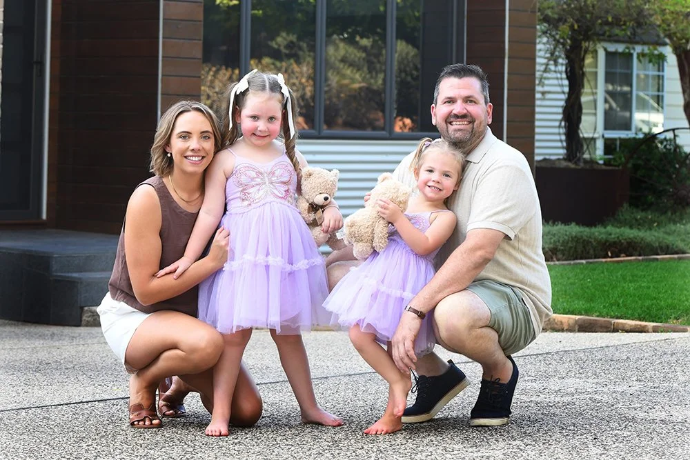 A happy family of four posing outdoors on a sidewalk in front of a modern house. The mother and father kneel on each side, each holding a young girl dressed in purple tutu dresses holding teddy bears.