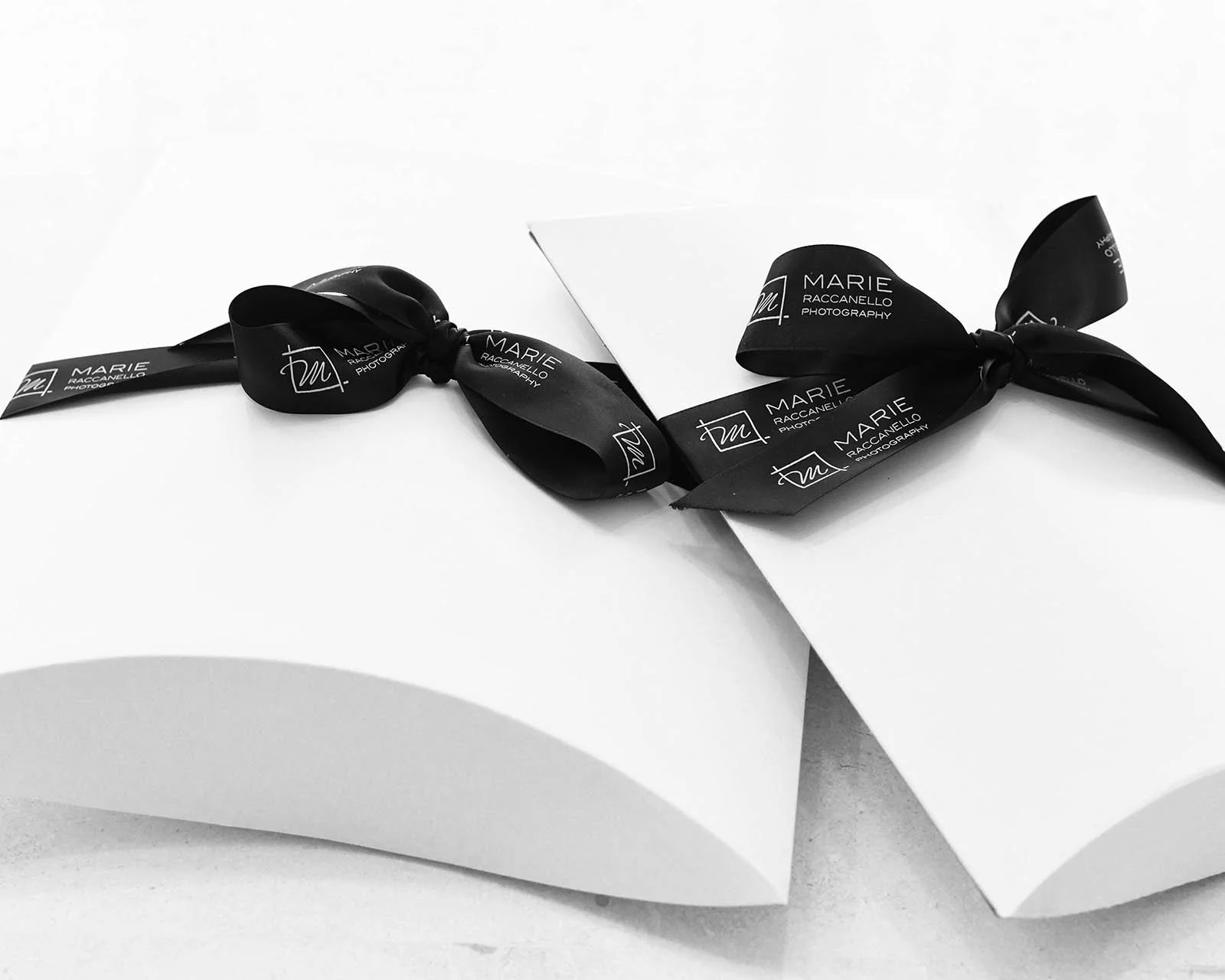 Black ribbon with white text and logo, tied around a white box, with another box partially visible beneath, on a light surface.