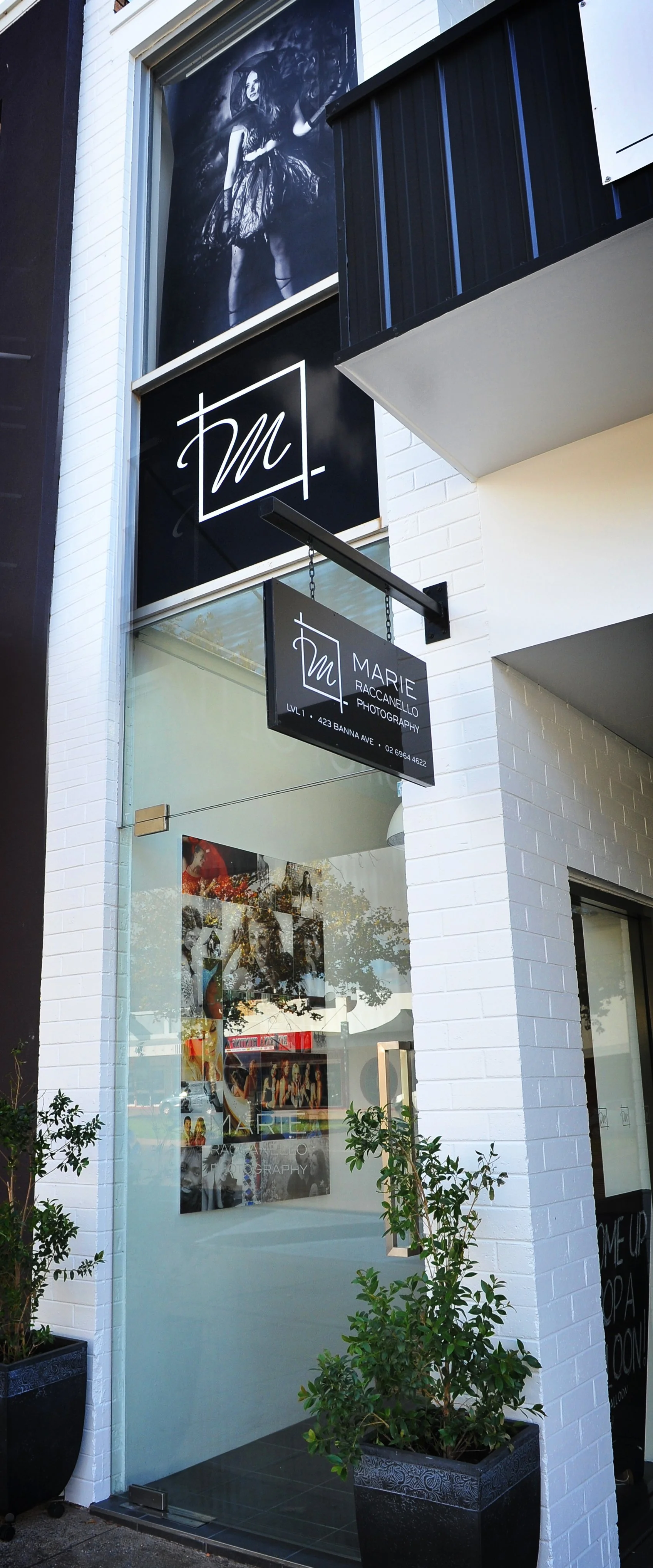 Front view of a photography studio storefront with signage. The sign reads 'Marie Raccanelo Photography' with contact details, and a large black-and-white portrait of a woman in a dress is displayed above. Inside, framed photos are visible through the glass.