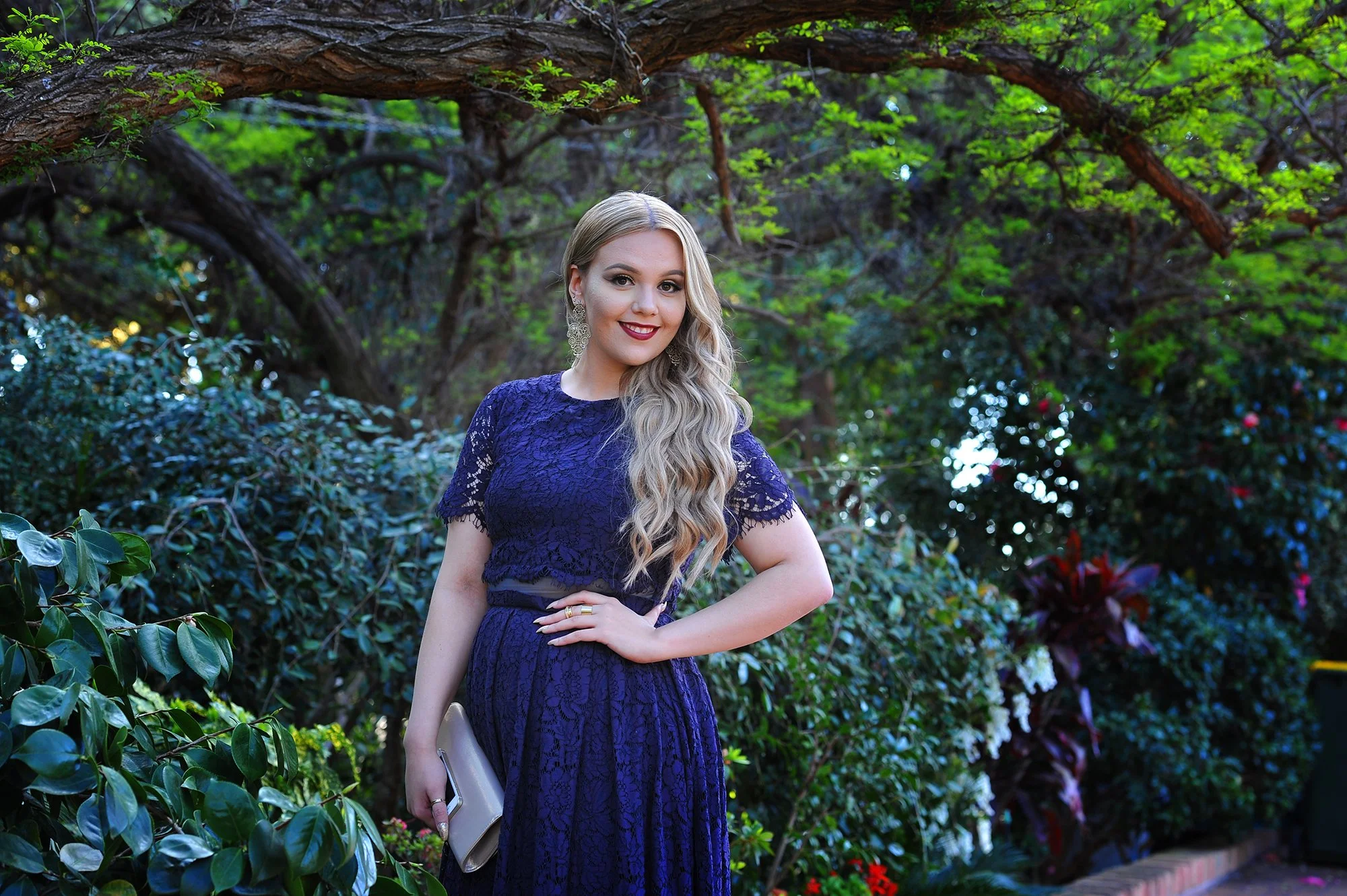 A young woman with long blonde hair wearing a dark purple lace dress, holding a beige clutch and gold-colored phone, standing outdoors in a garden with lush green foliage and trees.