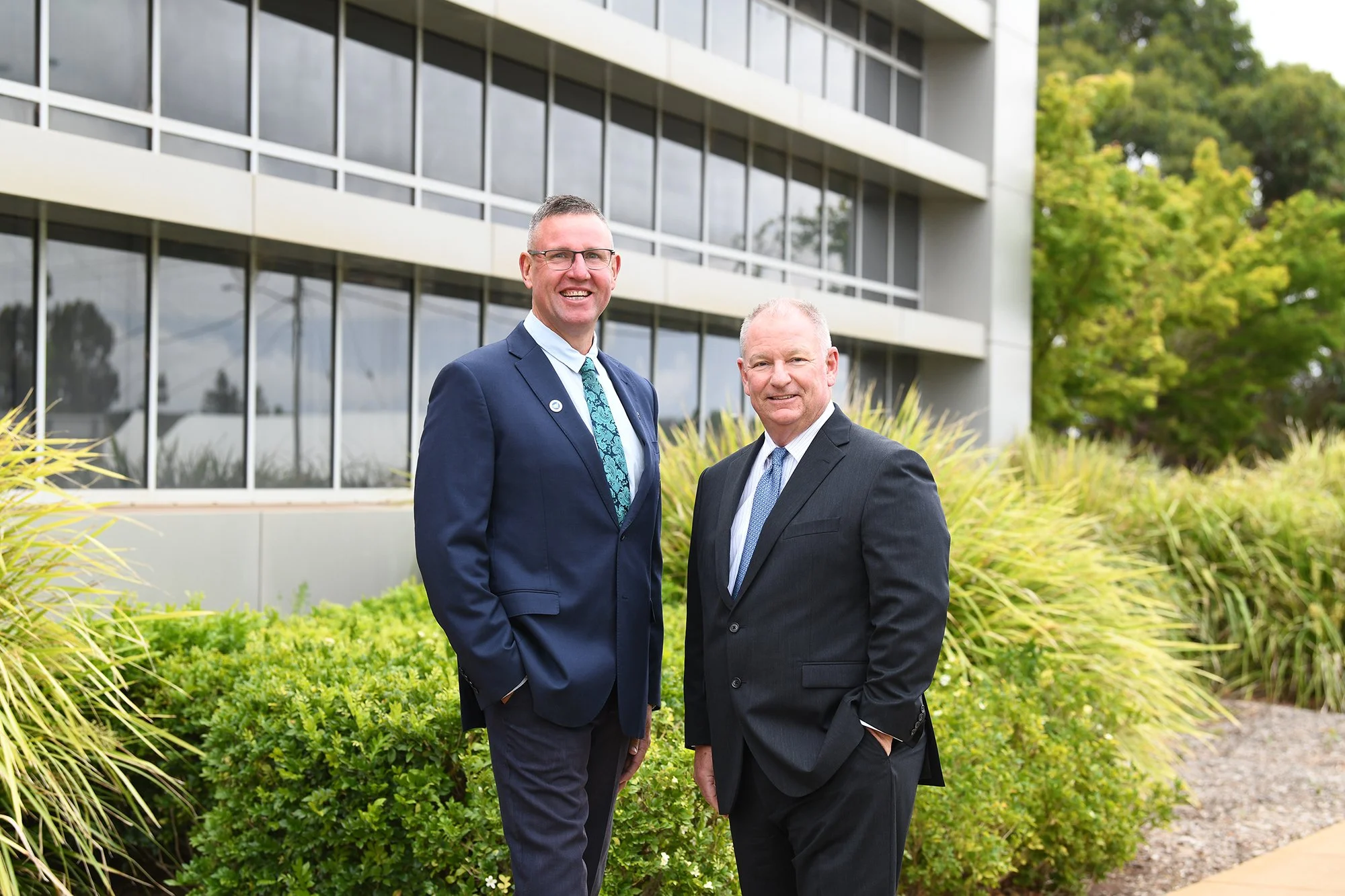 Two men in suits standing outdoors in front of a modern building with large glass windows, surrounded by green plants and trees.