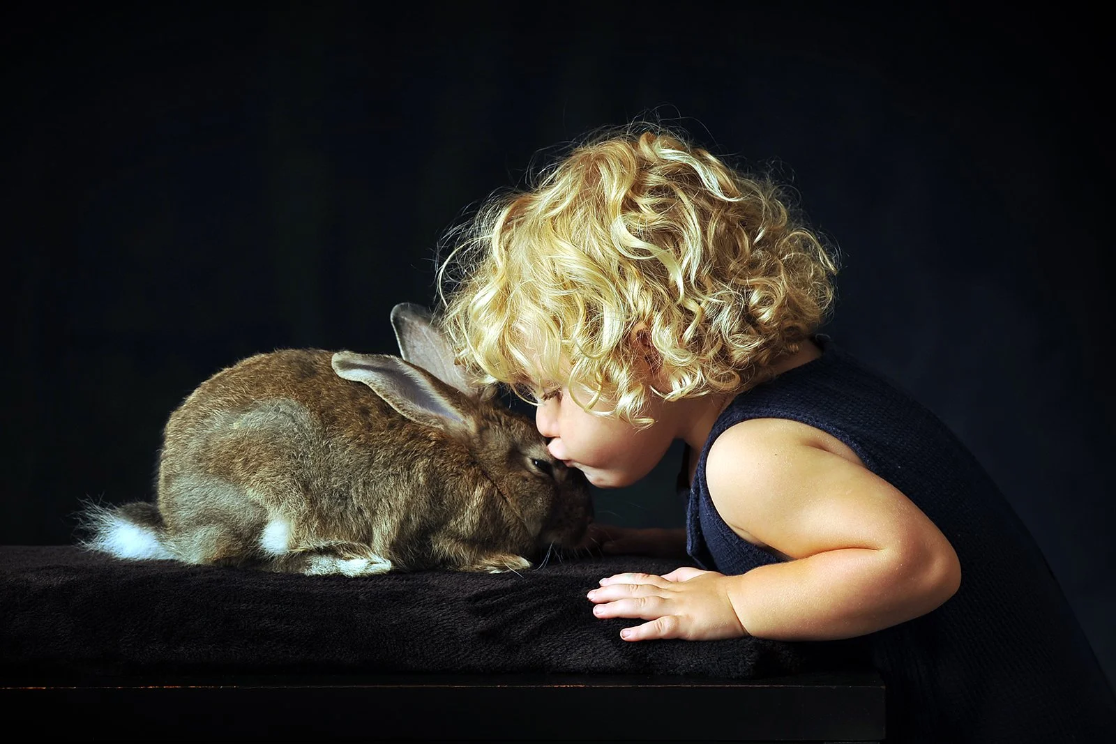 A young child with curly blonde hair gently touching foreheads with a brown rabbit against a dark background.