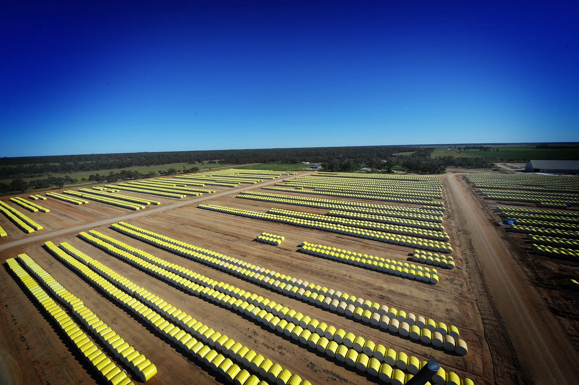 Aerial view of a large solar farm with numerous yellow solar panels arranged in rows on a dirt field, with a forest and farmland in the background under a clear blue sky.