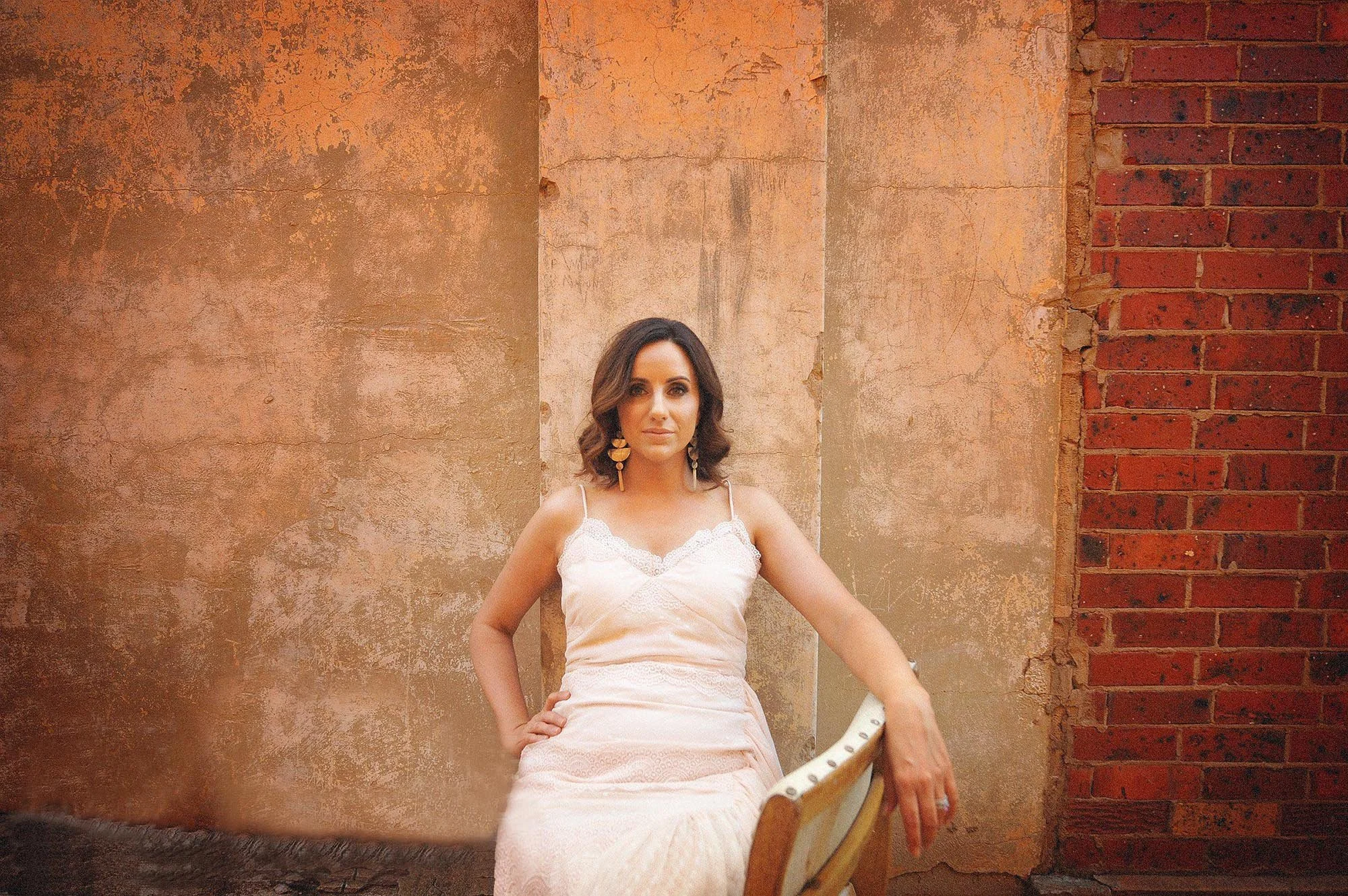 A woman with dark, wavy hair wearing a white dress with spaghetti straps and earrings, sitting on a vintage chair with a curved armrest against a textured, weathered wall with brick accents.