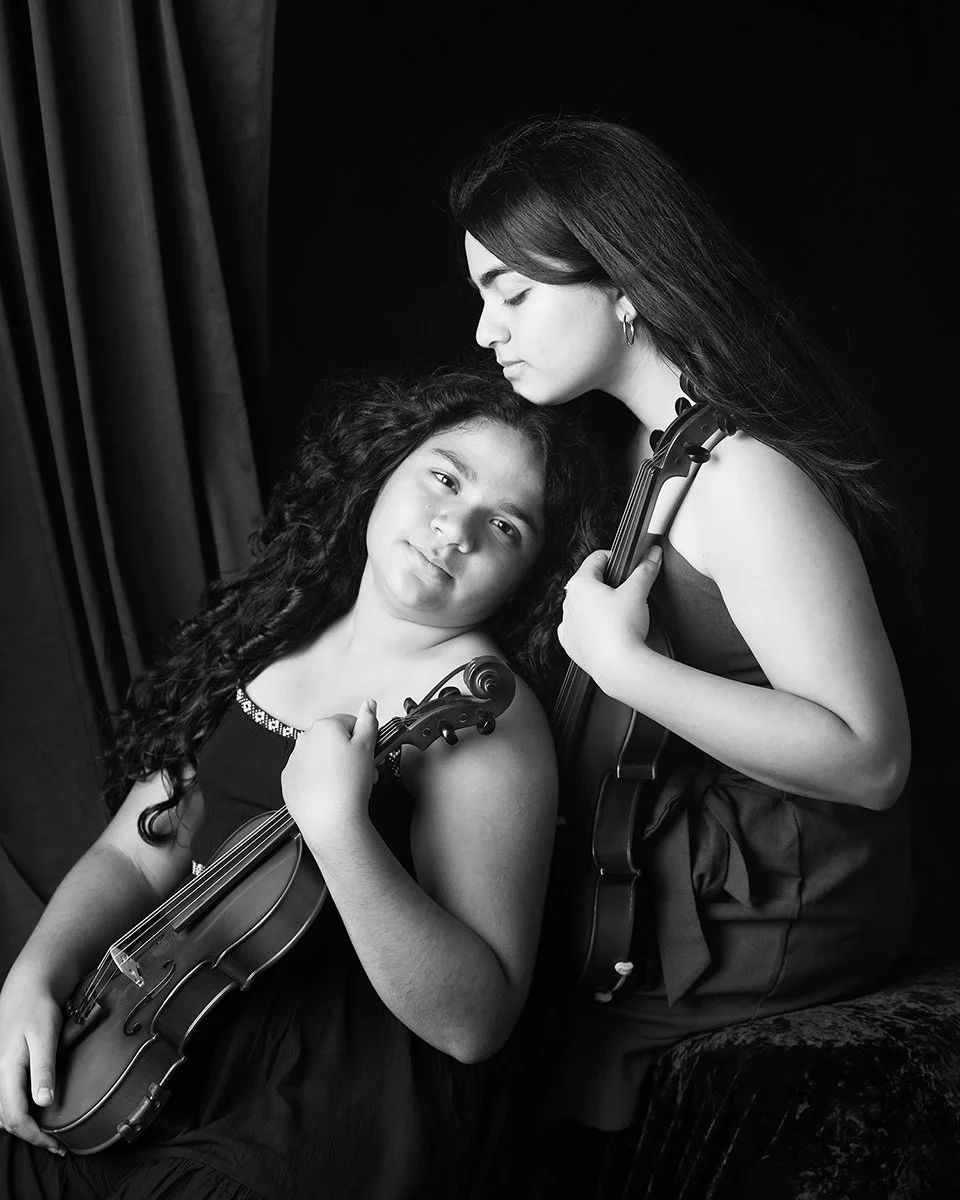 Two young girls with violins, one with curly hair leaning against the other with straight hair, against a dark background.
