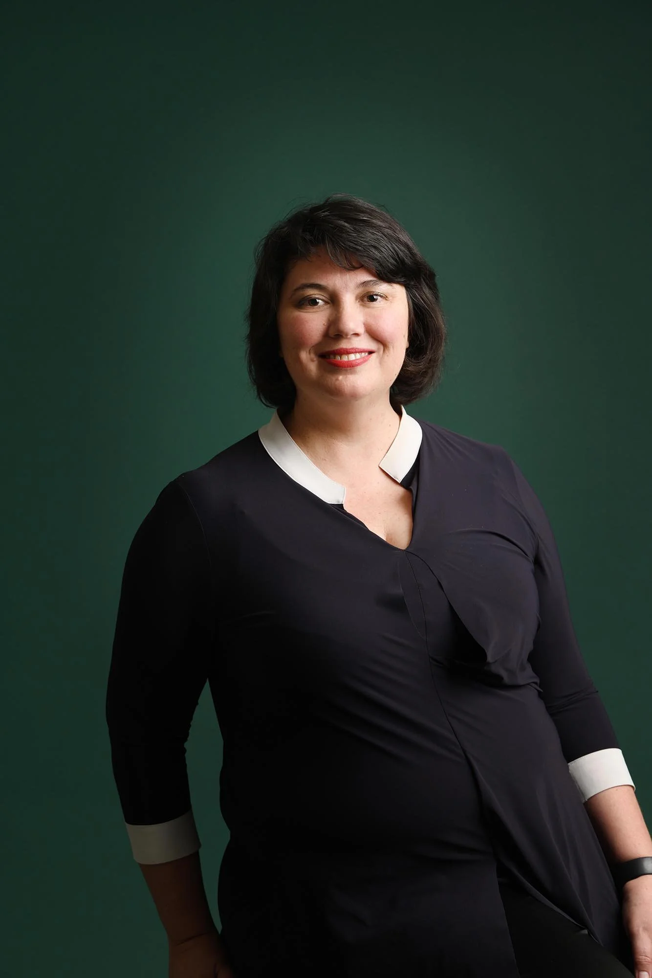 Portrait of a woman with dark hair wearing a dark dress with white cuffs, standing against a green background.
