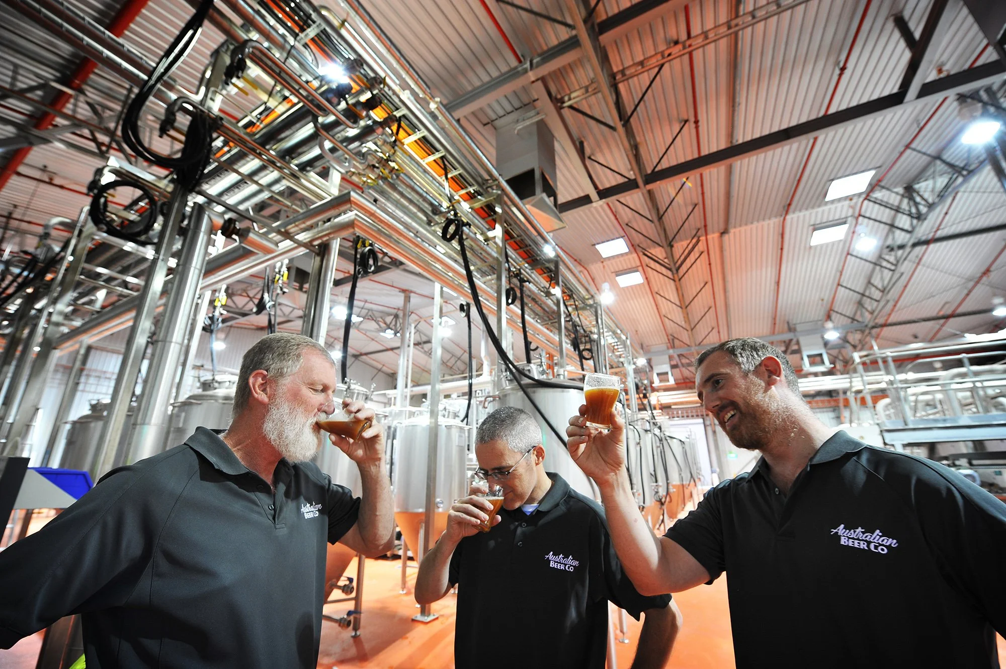 Three men in black shirts with 'Australian Beer Co' logo are drinking beer in an industrial brewery, with metallic pipes and manned tanks in the background.