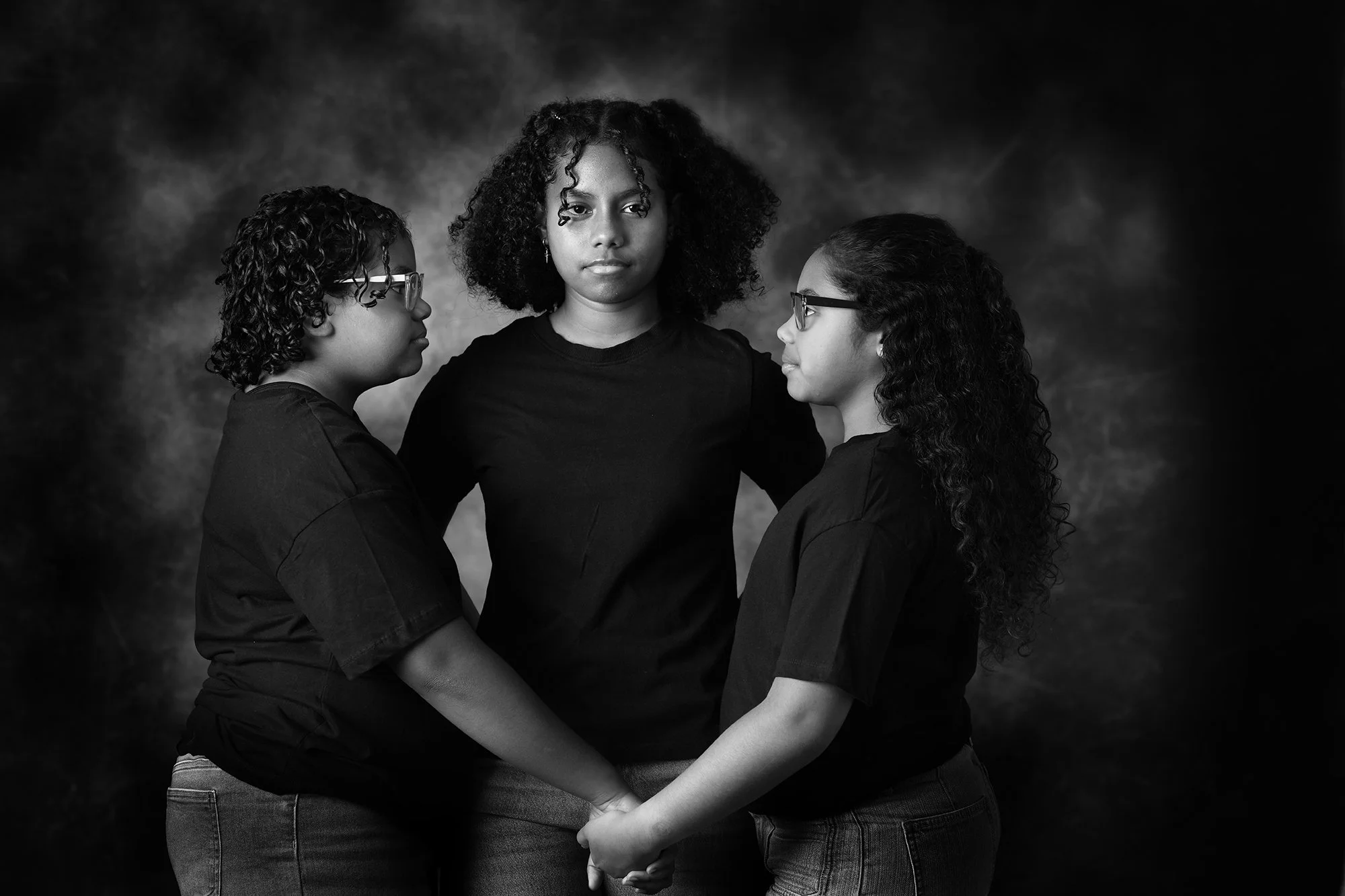 Three young women with curly hair and glasses standing close together, holding hands, in a black and white portrait against a dark background.