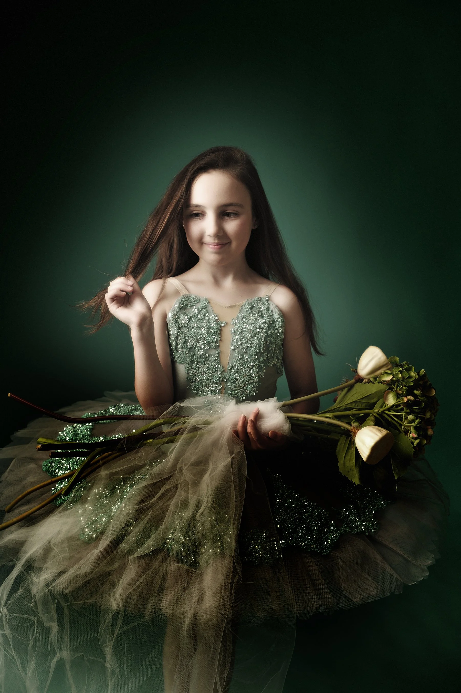 Young woman with long brown hair wearing a green embellished dress, sitting with a bouquet of white roses and green foliage, against a dark green background.