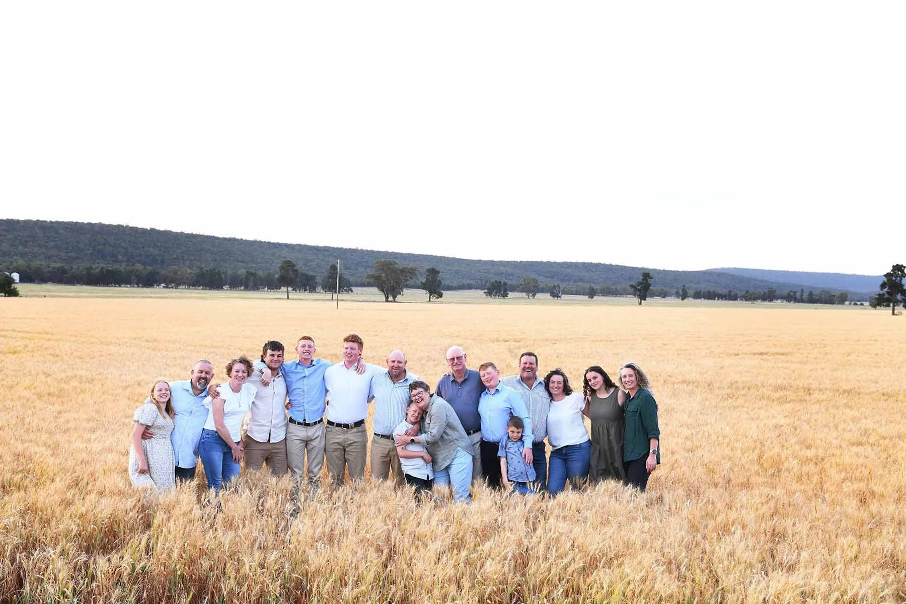 A large group of people, including men, women, and children, standing and smiling in a golden wheat field during daytime with distant hills and scattered trees in the background.