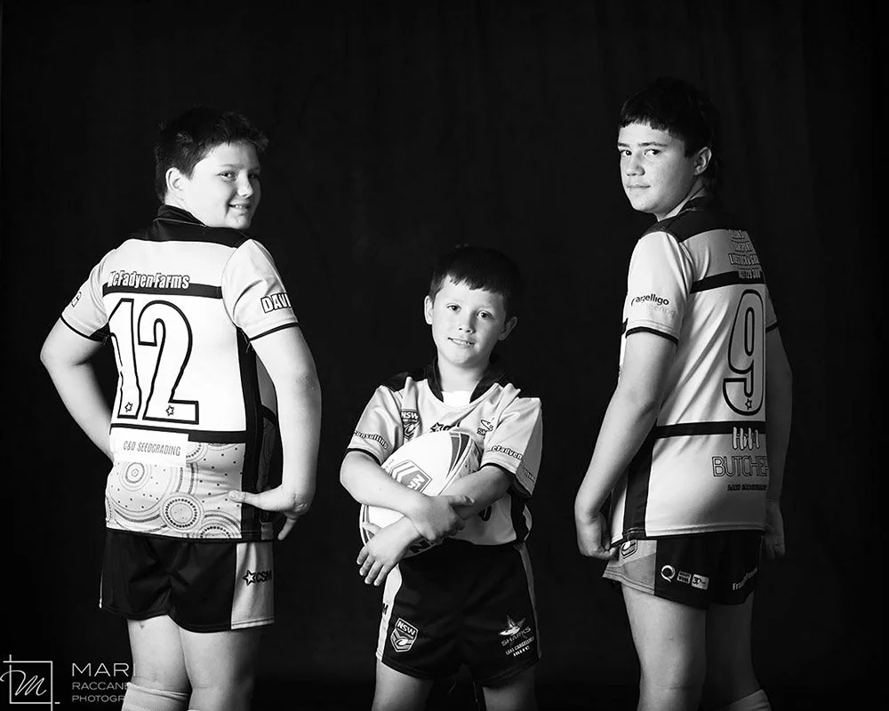 Three boys in rugby uniforms, one holding a rugby ball, posing against a dark background.