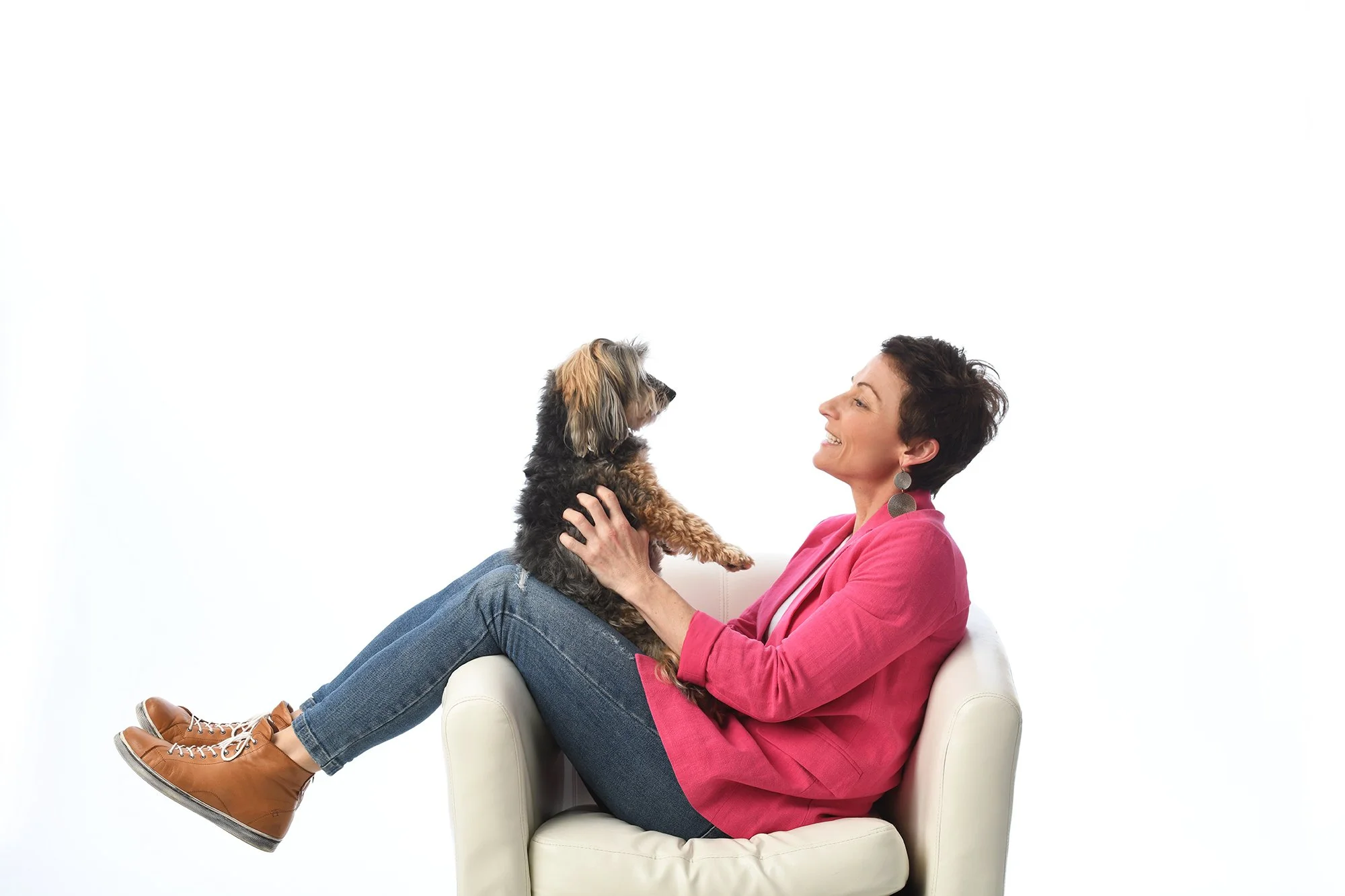 Woman in a pink blazer and jeans sitting on a white armchair, smiling and holding a small dog in her arms. The dog is looking at her.
