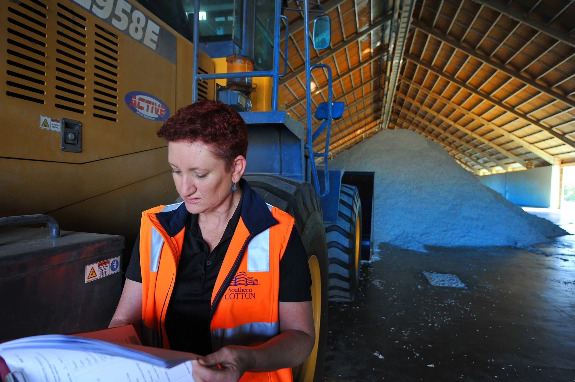 A woman with short red hair wearing an orange safety vest with 'Southern Cotton' logo, inspecting documents in a warehouse filled with cotton and a large yellow industrial machine.