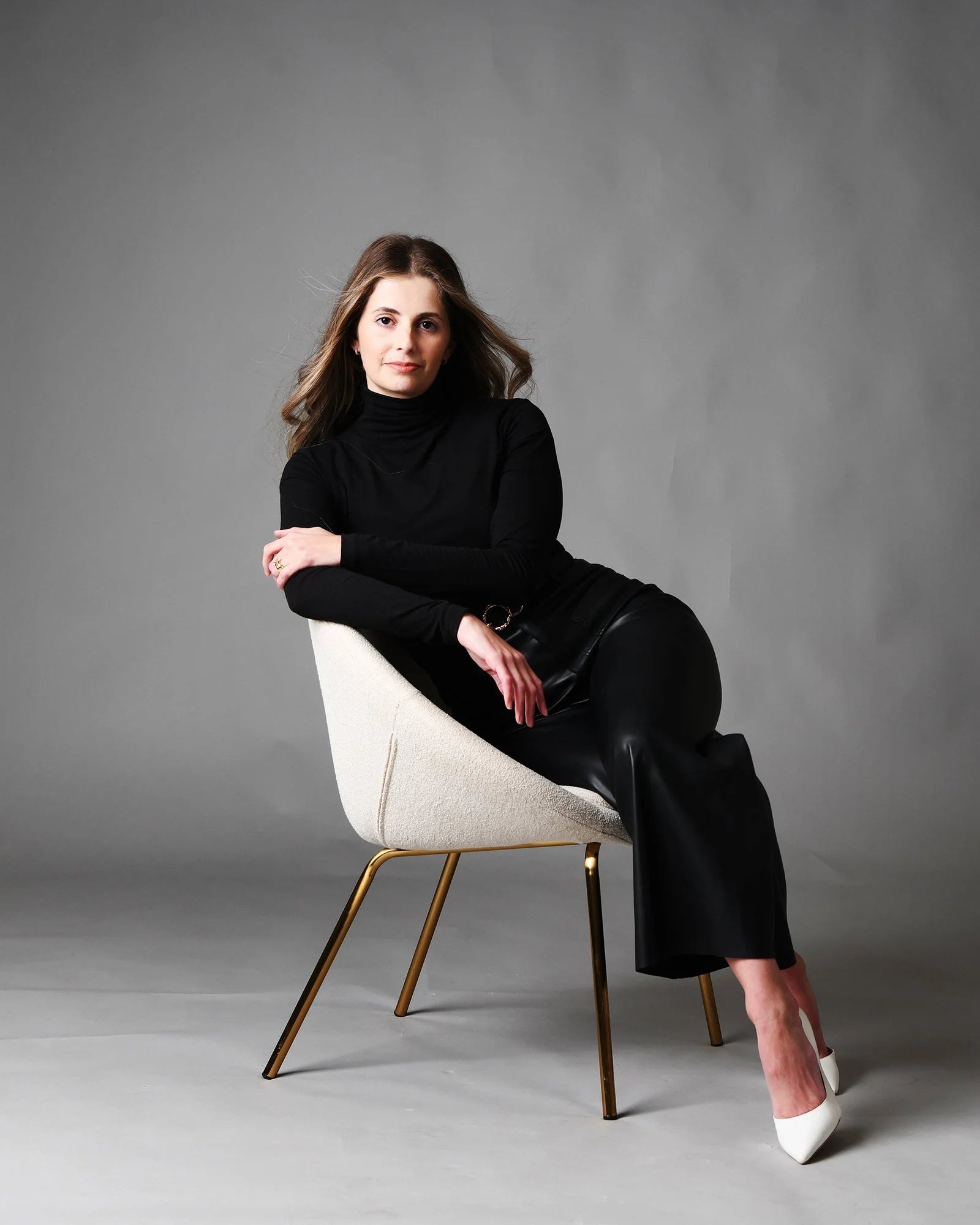 A woman with long brown hair wearing a black turtleneck and black pants, sitting on a white chair with gold legs against a gray background. She is leaning sideways with her left arm resting on the chair and her right arm crossed over her knees, weari