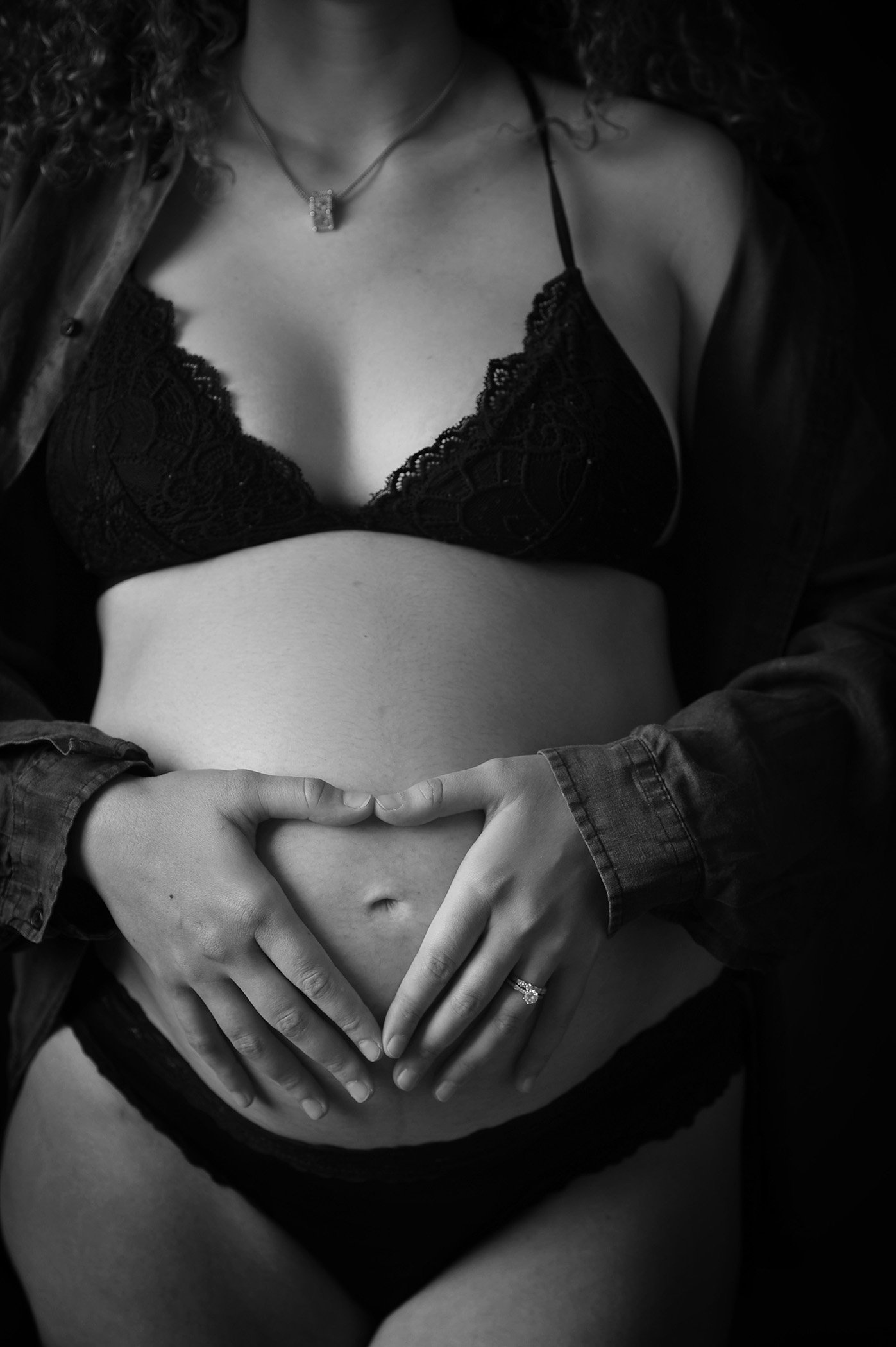 A woman wearing a black lace bra and a necklace, with her hands formed into a heart shape on her abdomen, in a black and white photograph.
