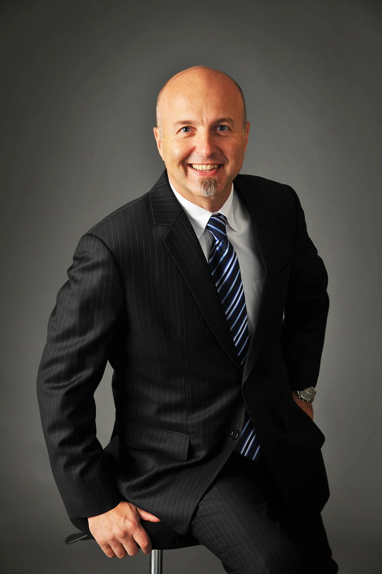 A man in a dark pinstripe suit with a blue and white striped tie, sitting on a stool against a plain dark background, smiling at the camera.