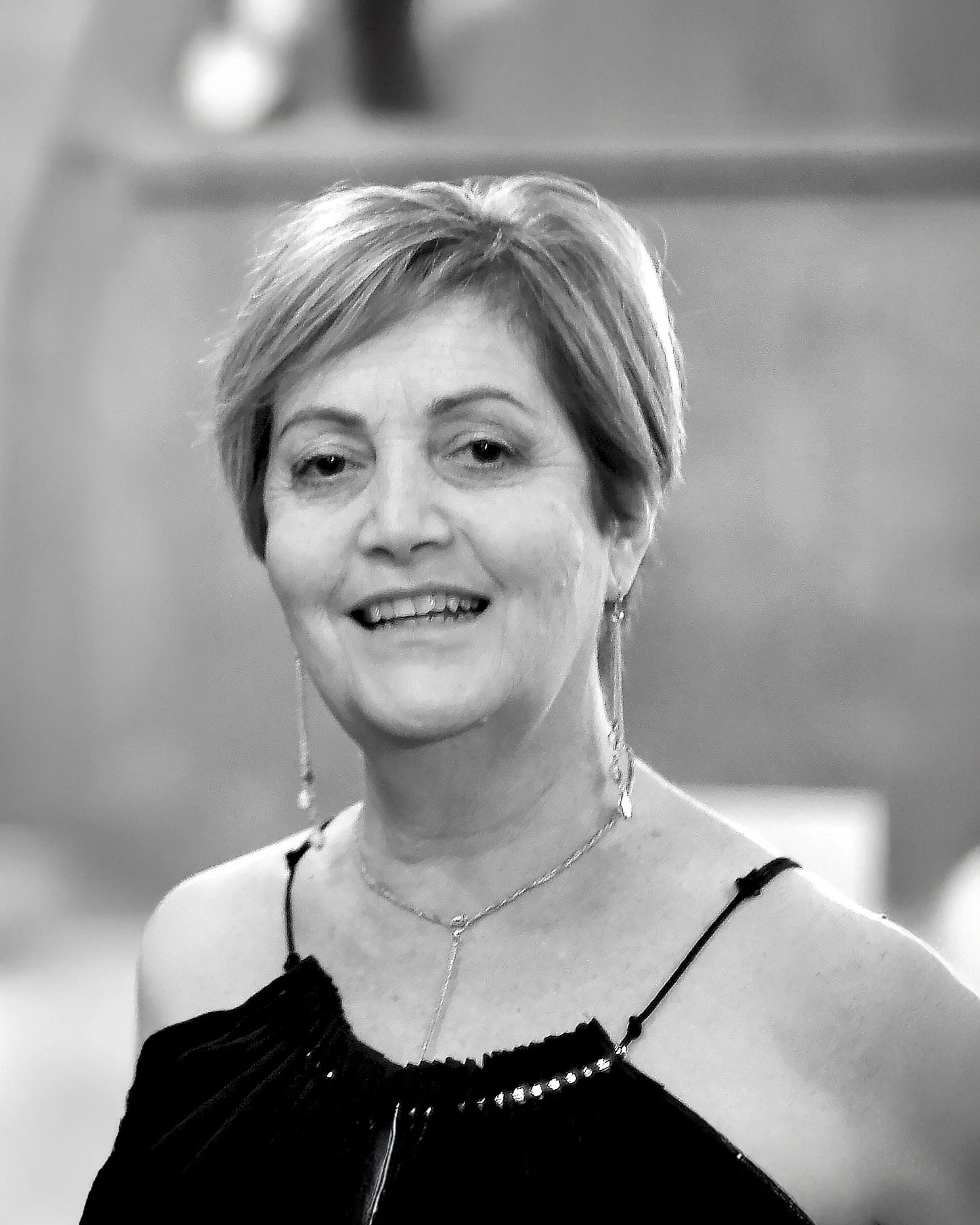 Black and white photo of a smiling woman with short hair, wearing a spaghetti strap dress with a pearl detail, earrings, a necklace, and a pendant.