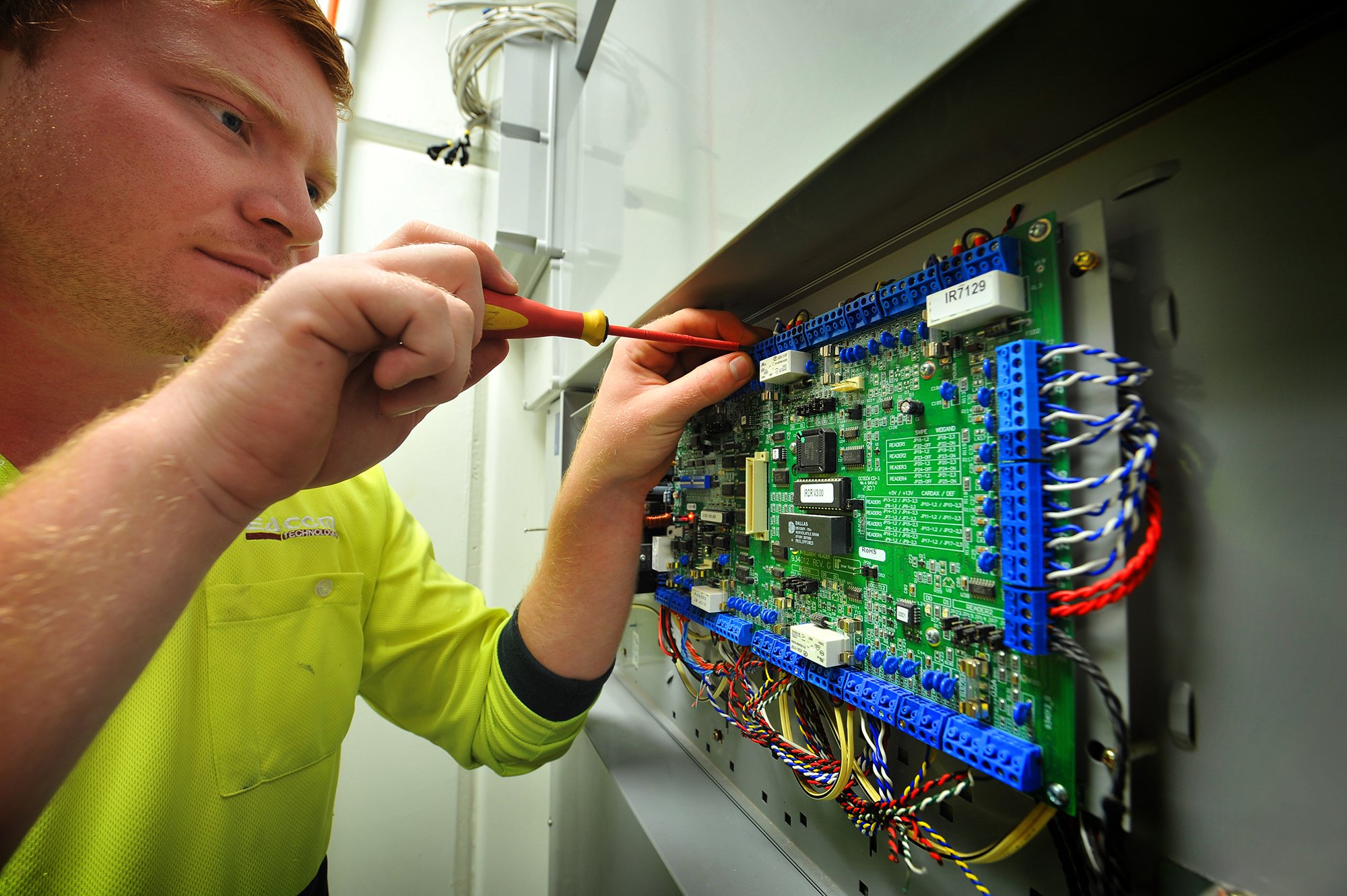 A technician in a yellow shirt working on an electronic control panel with circuit boards and wiring.