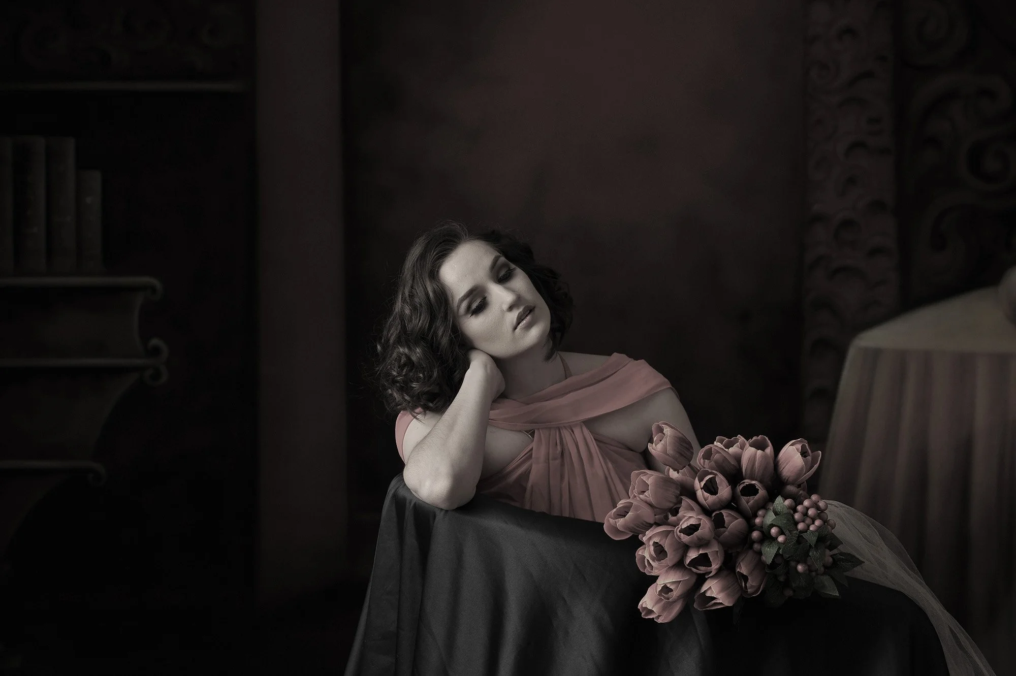 A woman with dark, curly hair is sitting at a table, resting her head on her hand and looking down thoughtfully. She is wearing a pink dress with a bow detail on the sleeve and there is a large bouquet of pink tulips in front of her. The background i