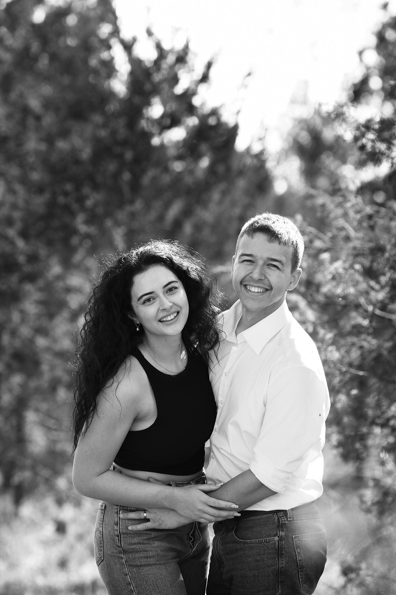 A black and white photo of a smiling young couple outdoors, standing close together with trees in the background.