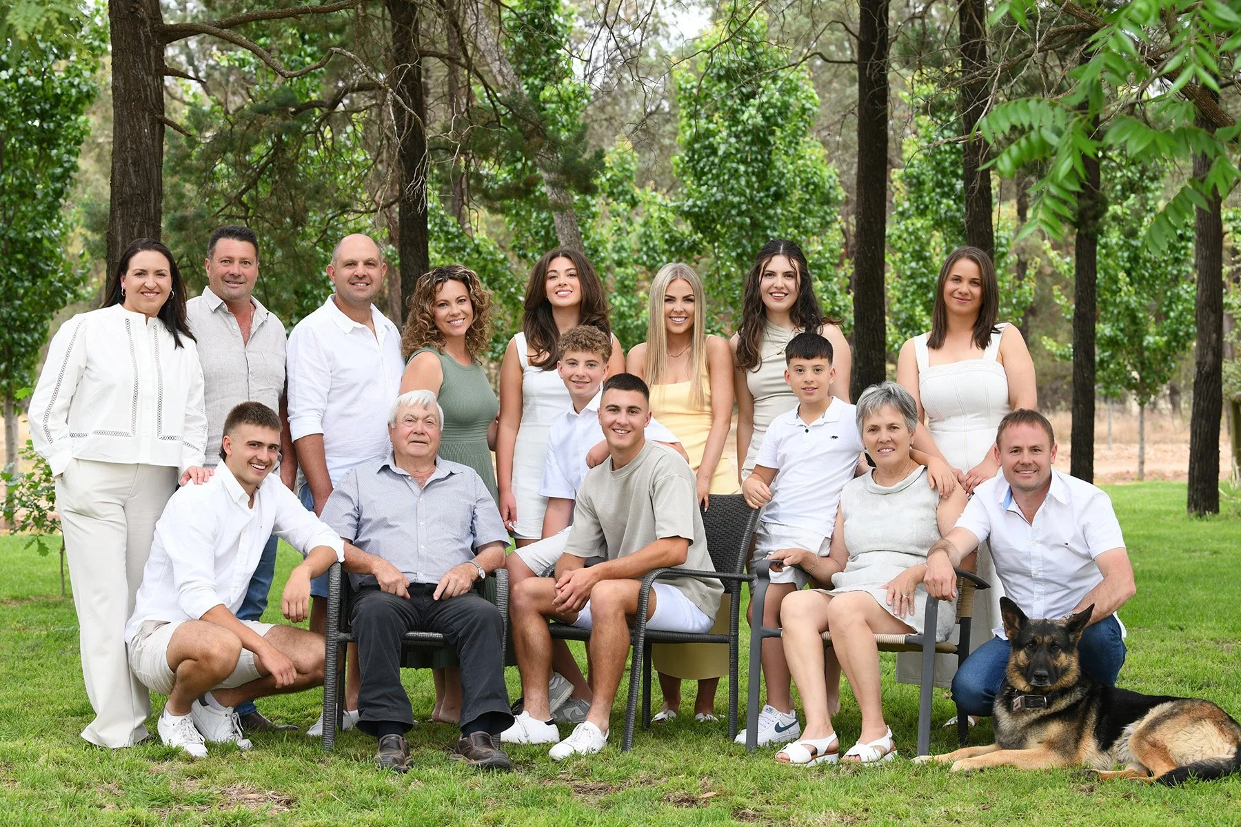 A large multigenerational family gathered outdoors in a wooded area, posing for a group photo. The family includes adults, teenagers, and elderly members, along with a German Shepherd dog lying on the grass. They are dressed in casual light clothing 