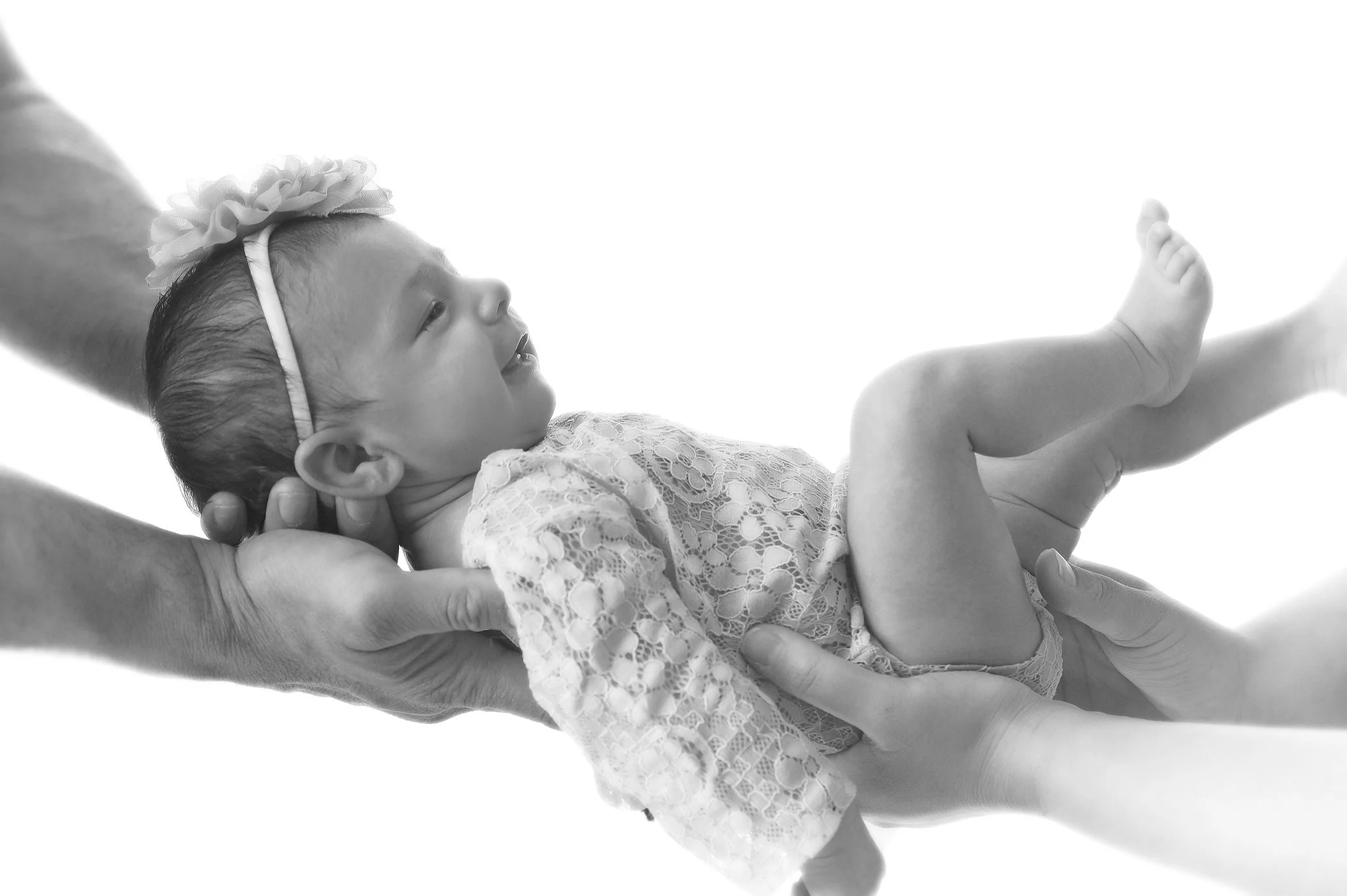 A baby girl in lace clothing and a flower headband being held and examined by a person's hand, with a focus on her face and body in black and white.