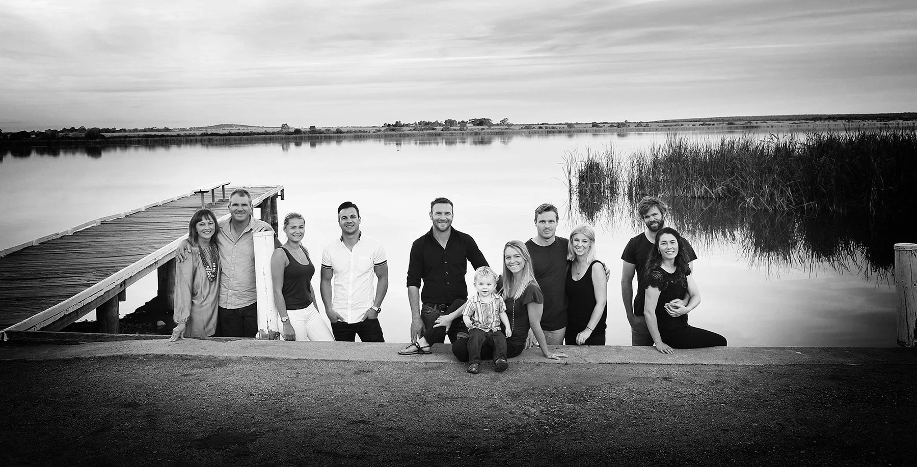 A group of twelve people, including children, posing together on a dock by a lake with calm water and reeds in the background, outdoors during the day.