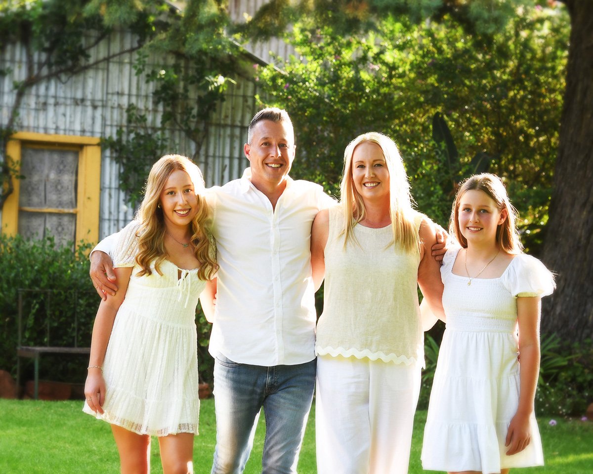 A family of four standing together outdoors, smiling, with a background of trees and a wooden building, all dressed in light-colored clothing.