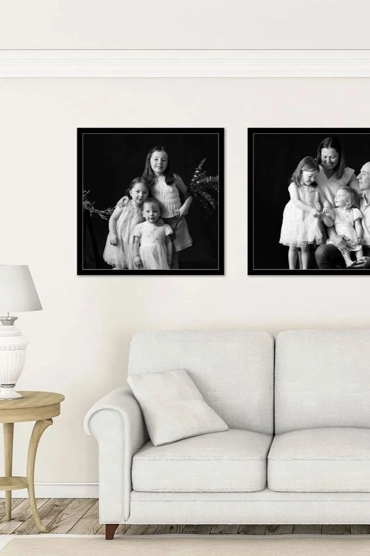 Two framed black and white family photographs hanging on a white wall above a white sofa with a cushion, next to a side table with a lamp.