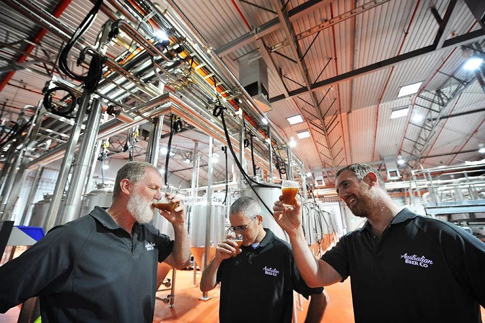 Three men are inspecting and tasting beer in a brewery with brewing tanks and pipes in the background.